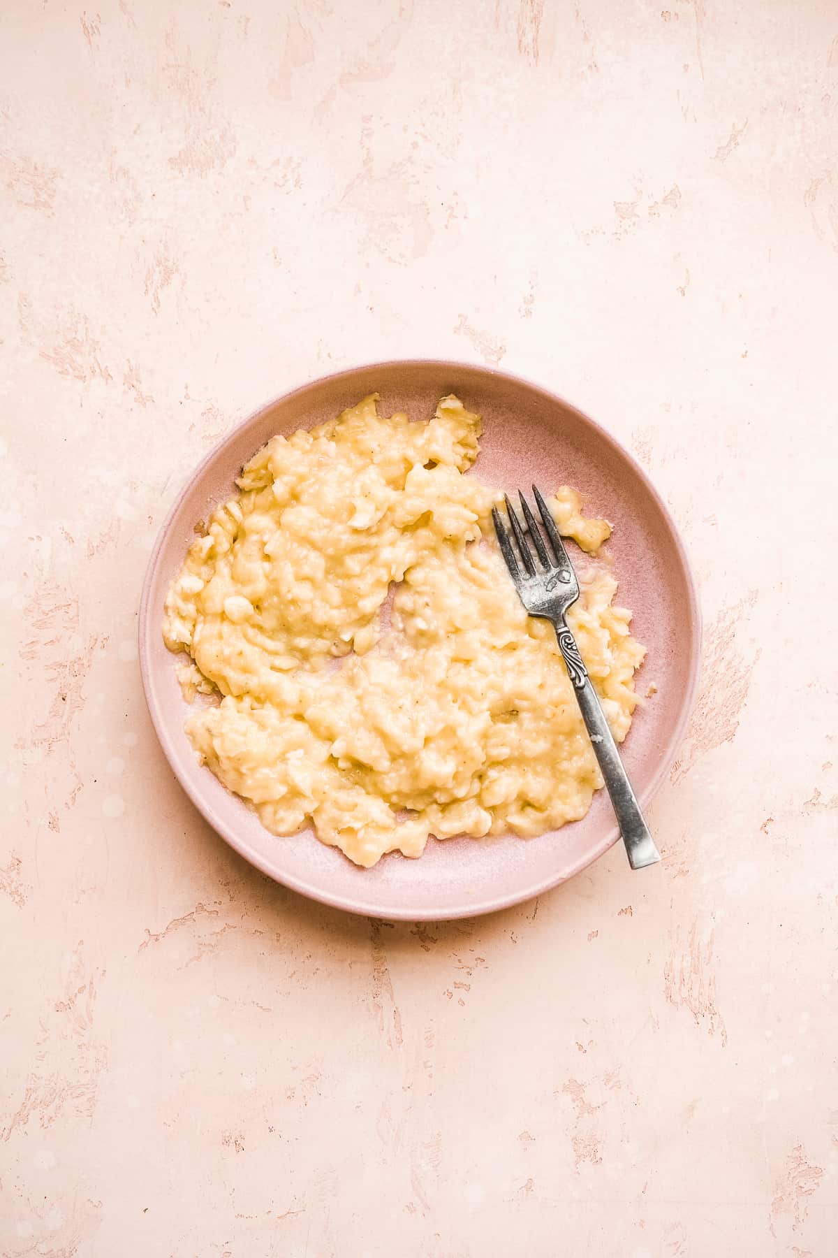 A pink plate with mashed bananas and a fork on the side.