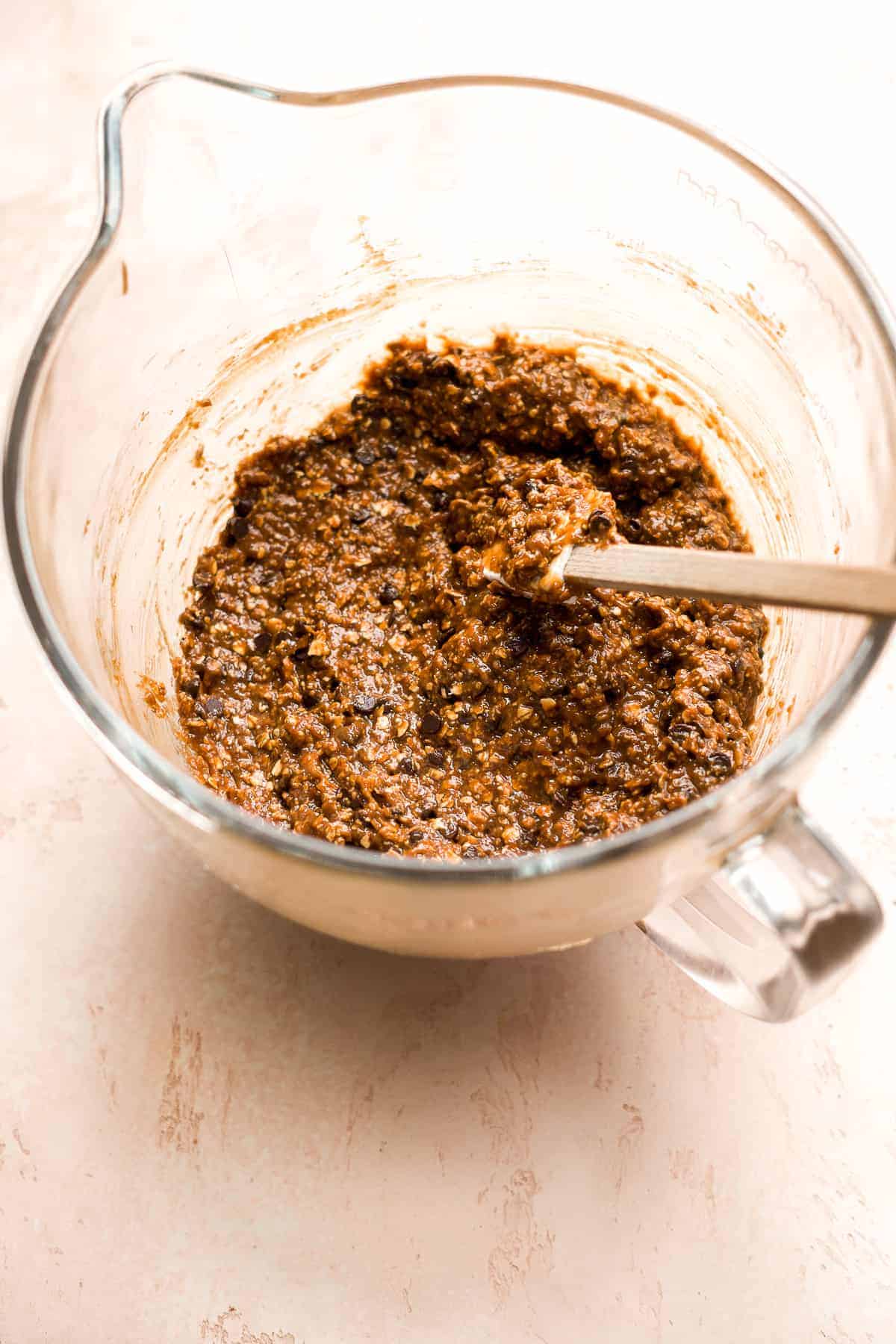 Glass mixing bowl with oatmeal chocolate chip cookie batter and a rubber spatula inside.