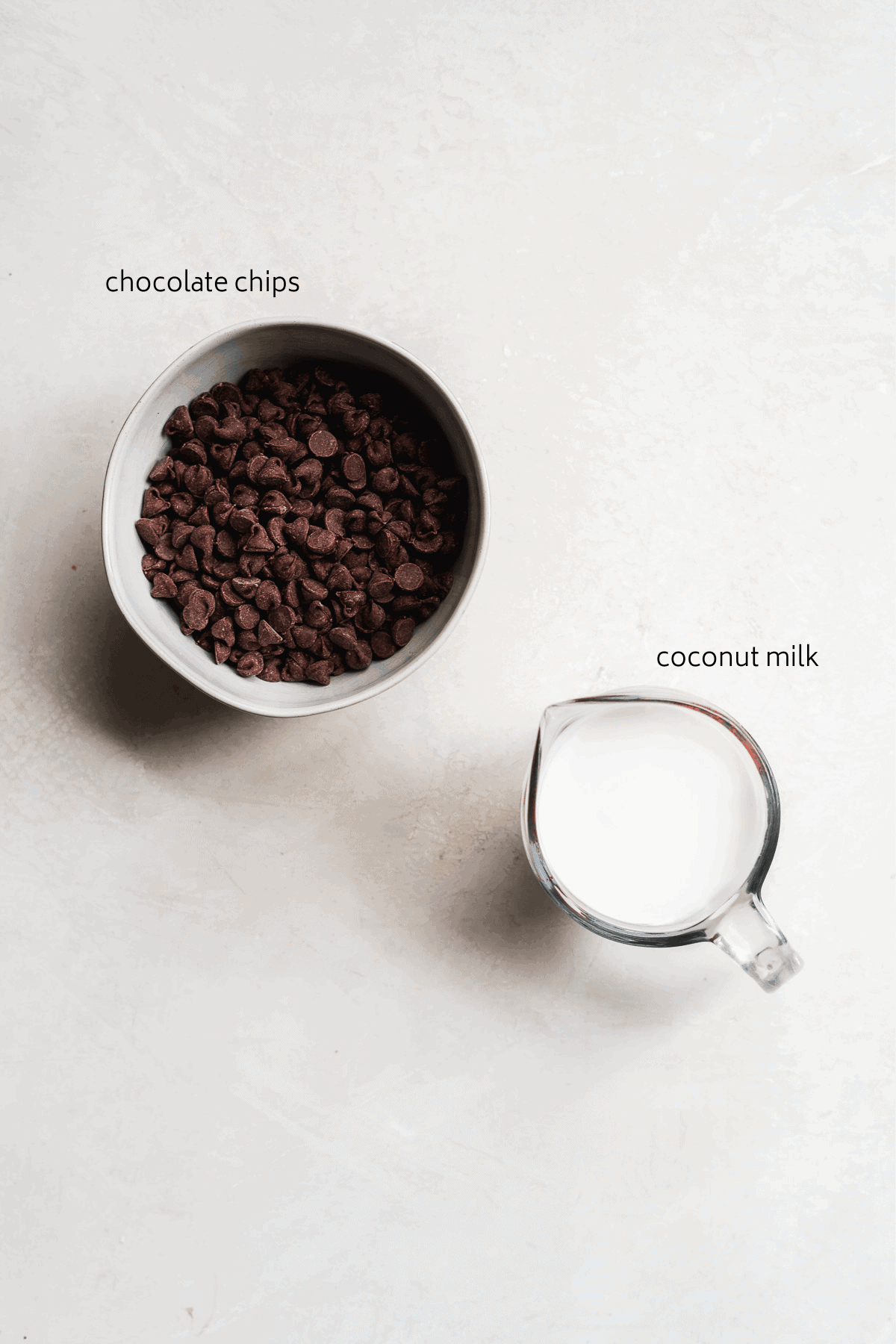 Bowl of chocolate chips and a measuring cup with milk on a white sufrace.