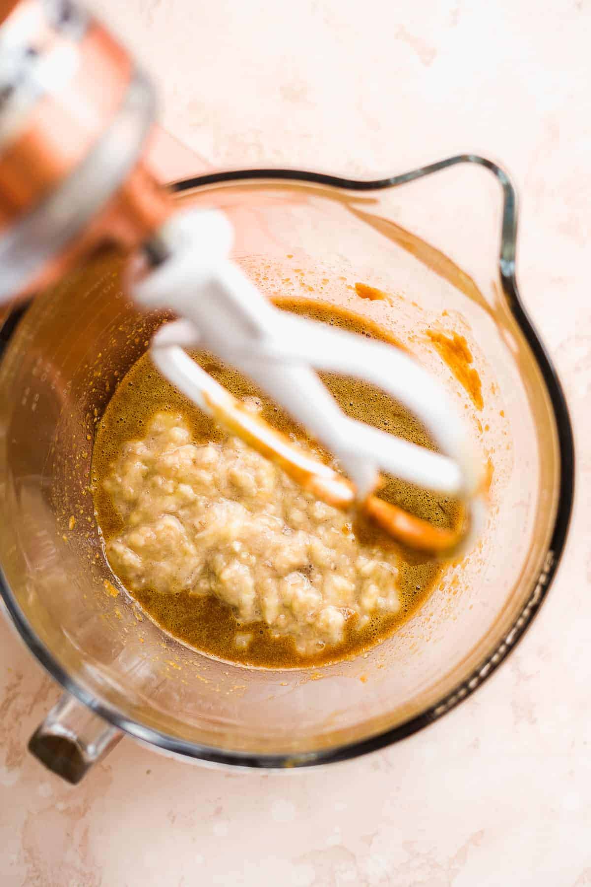 View inside a stand mixer with tan colored batter inside and mashed bananas.