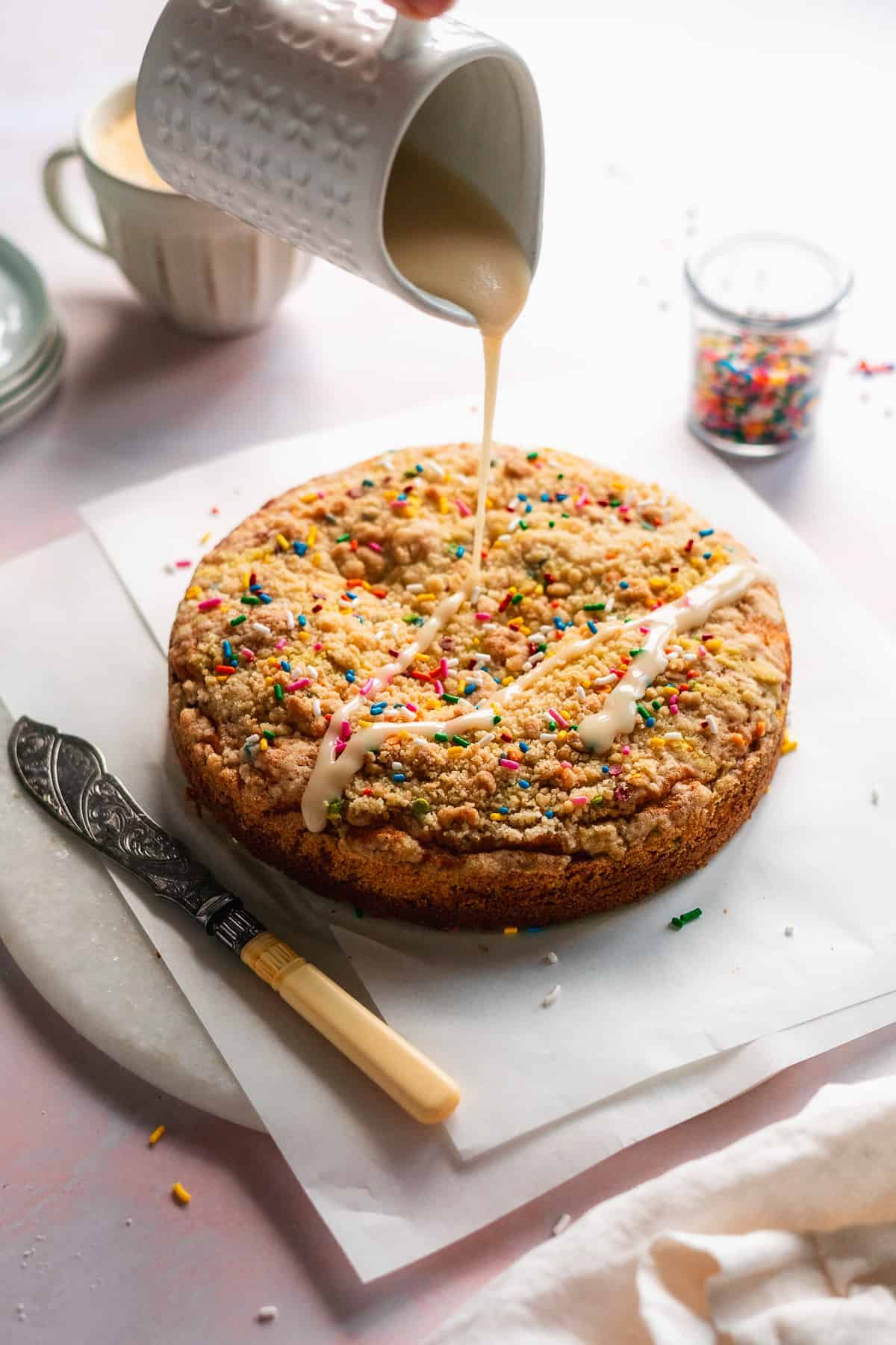 Hand pouring icing over top of a round coffee cake with sprinkles.