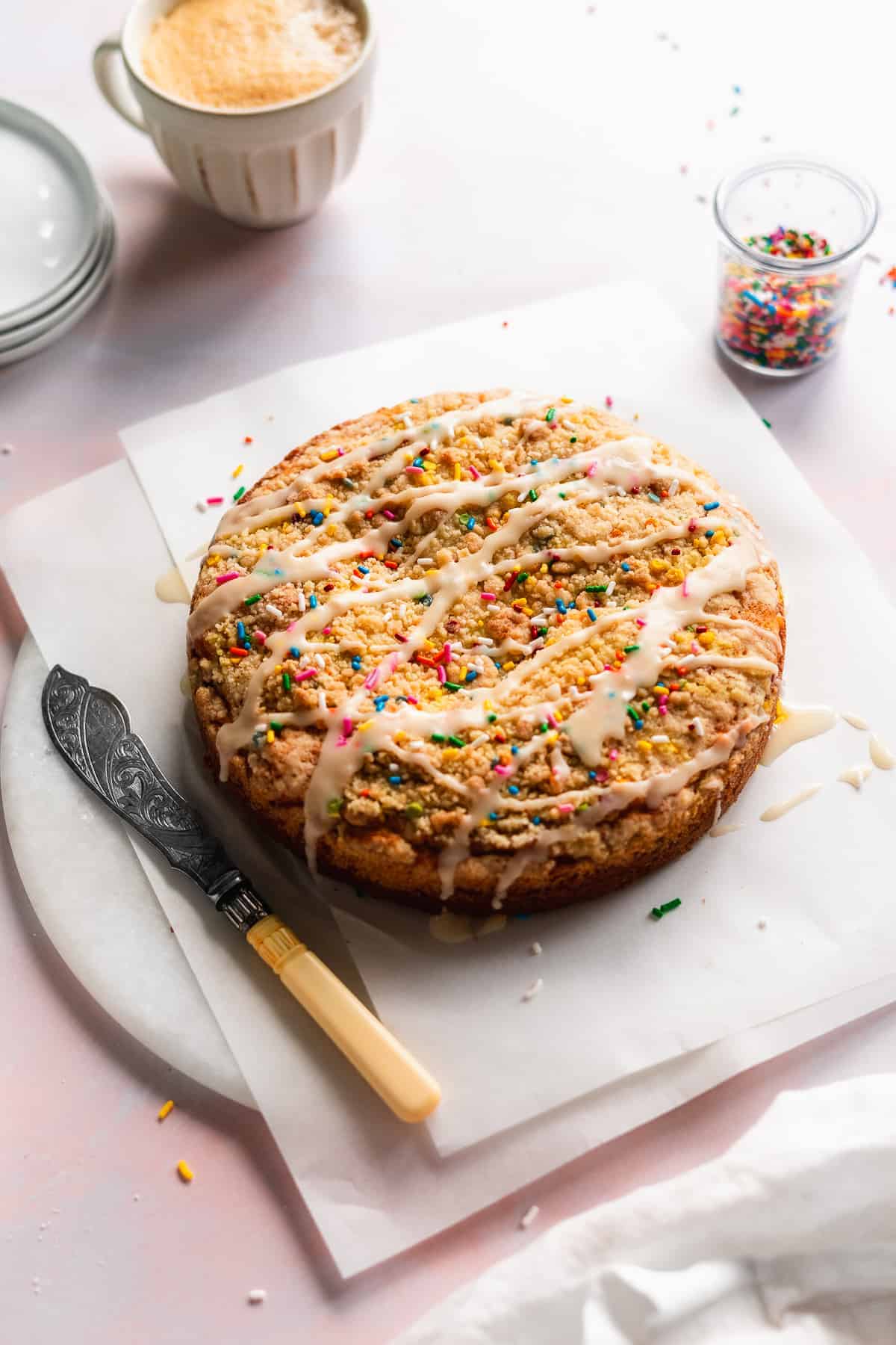 Circular coffee cake with sprinkles and icing on a white marble platter with a knife.