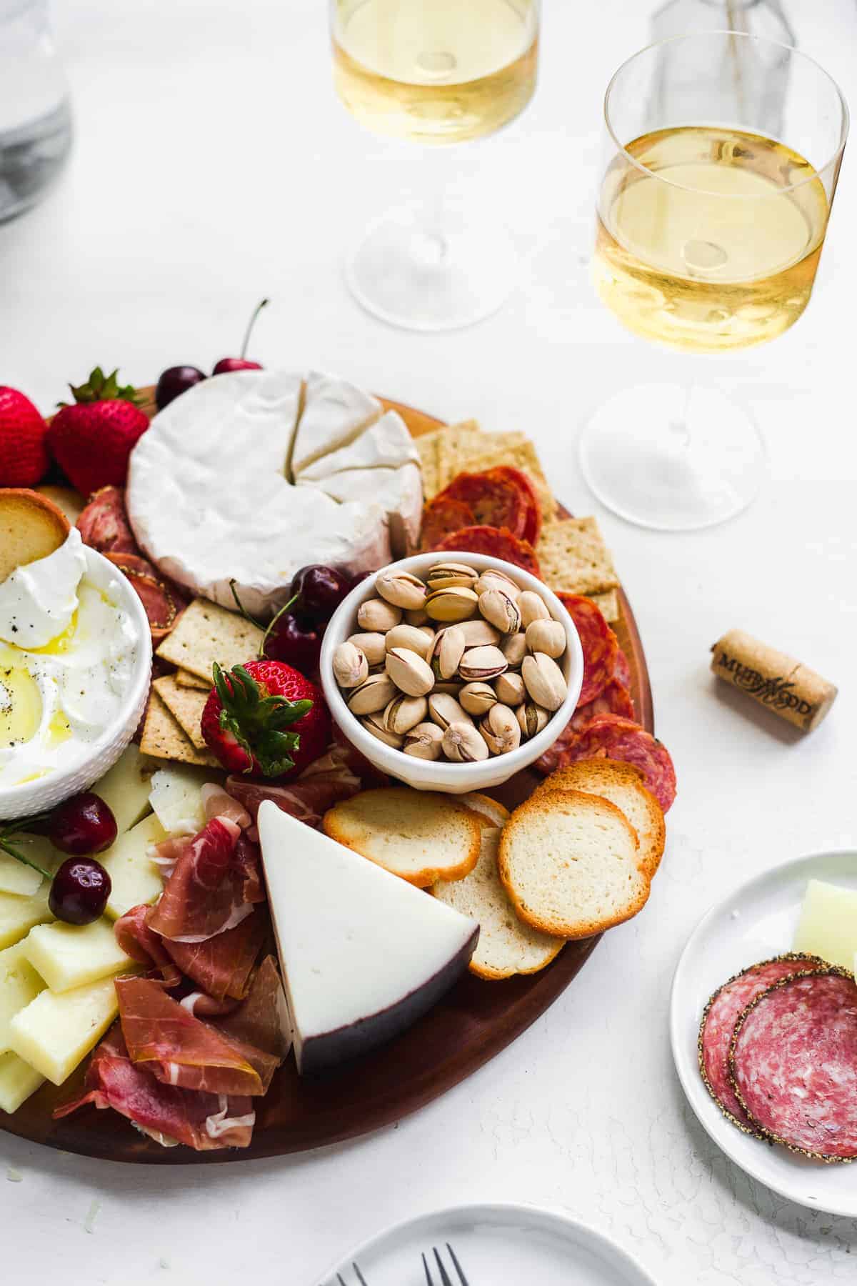 Charcuterie board with nuts and fruit and wine glasses in the background.