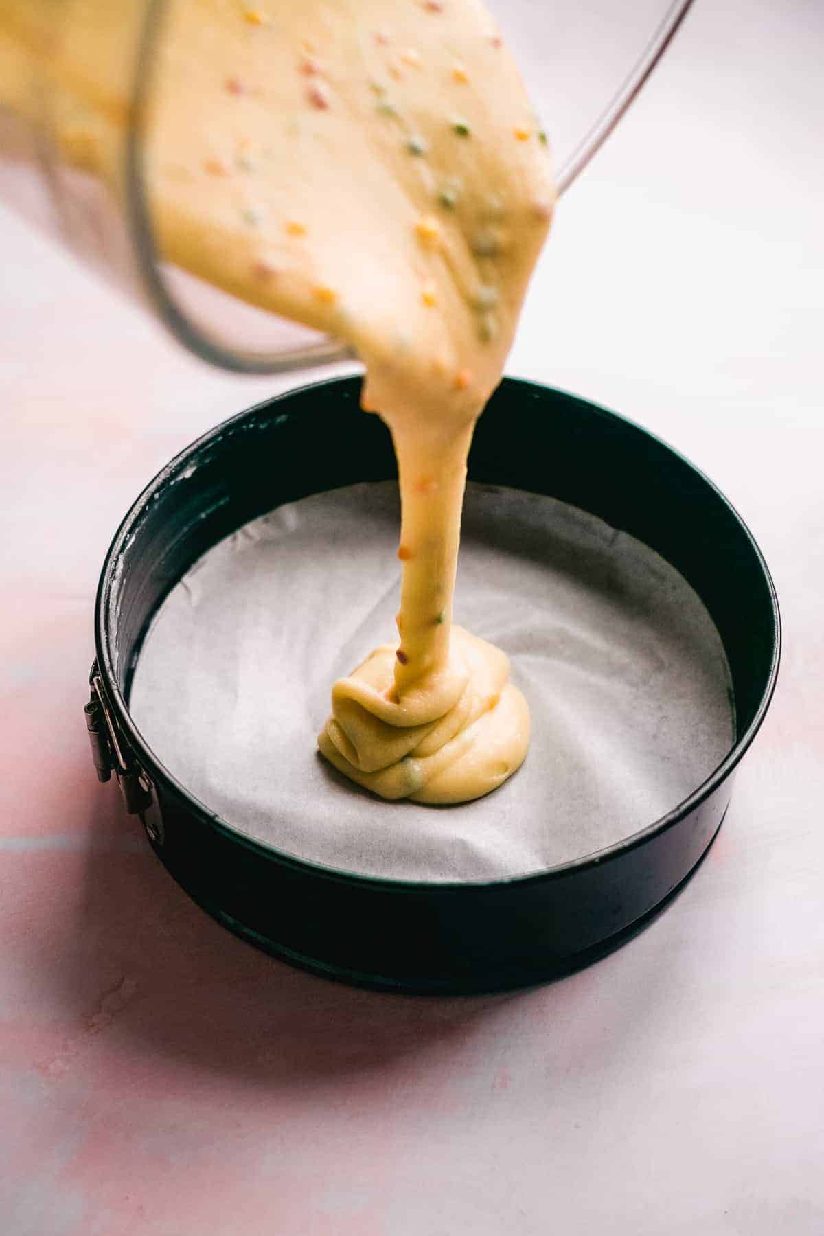 Cake batter with sprinkles being poured into a cake pan with parchment paper.