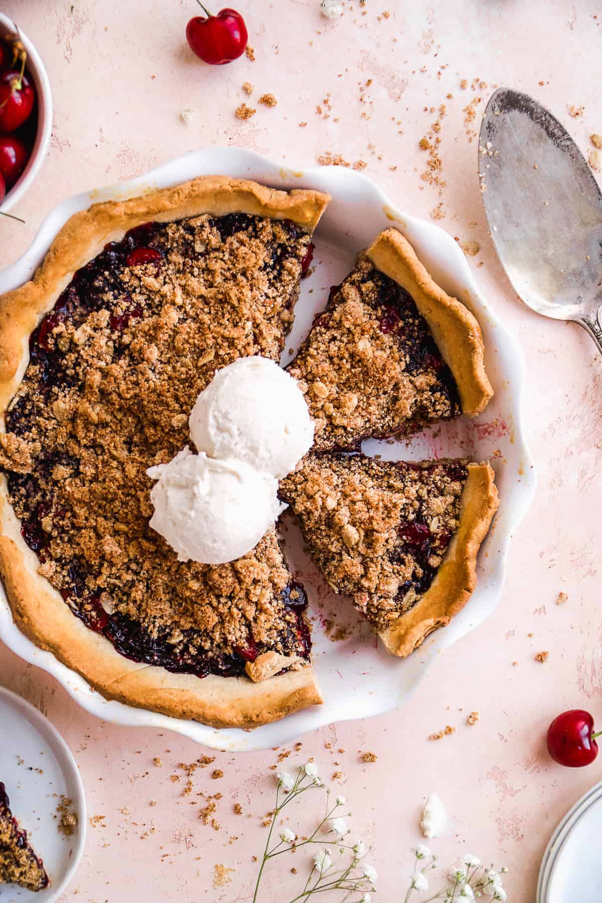 Cherry pie cut into slices with ice cream scoops in the middle on a pink surface.