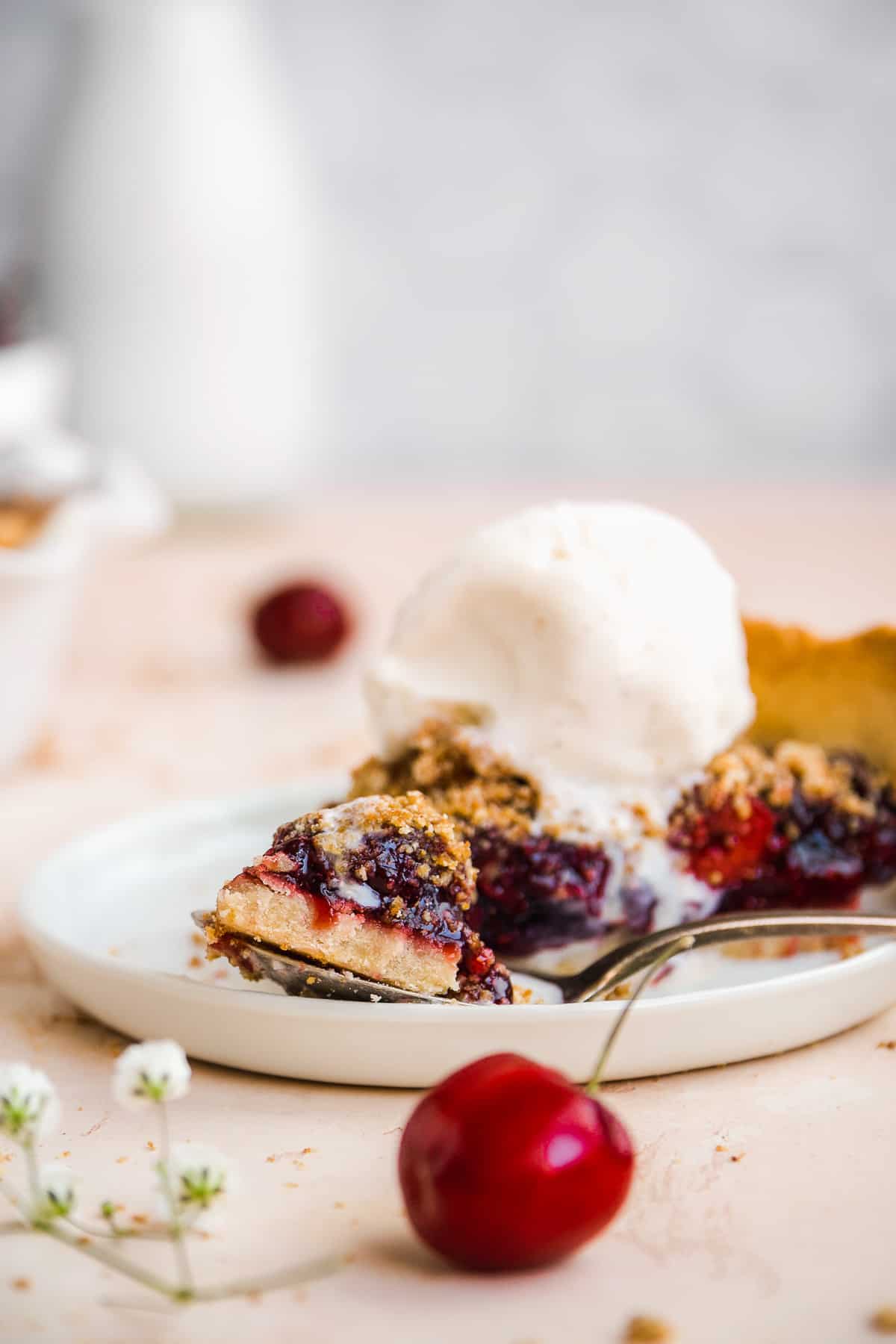 Slice of cherry pie on a white plate with a fork taking a bite.