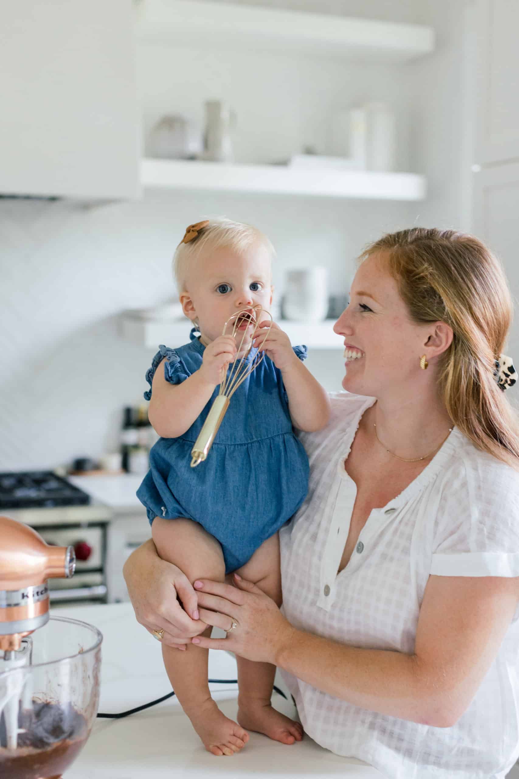 Woman holding baby on a counter with a whisk.