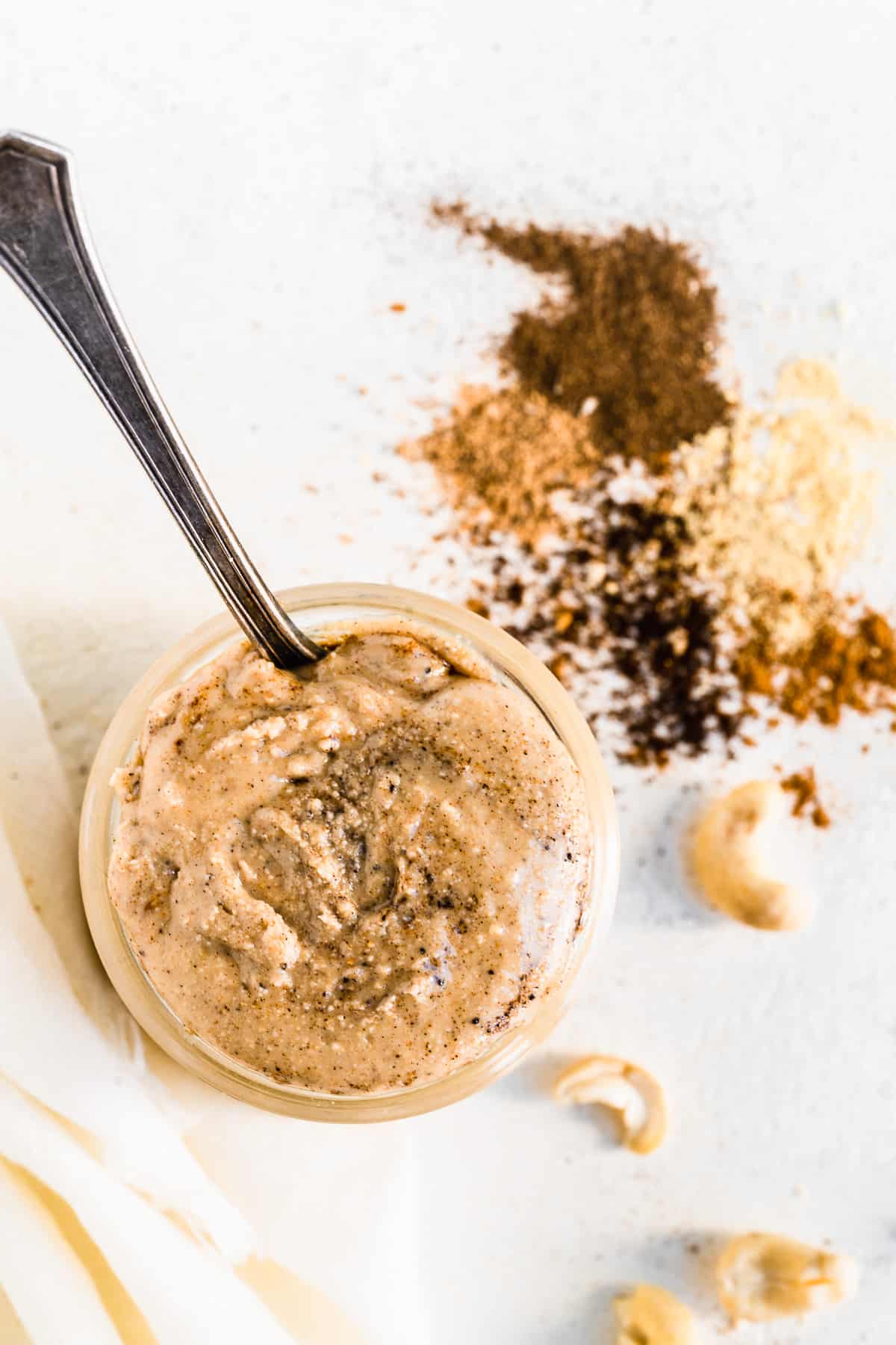 Jar of chia spiced cashew butter with a spoon inside and spices on the surface.