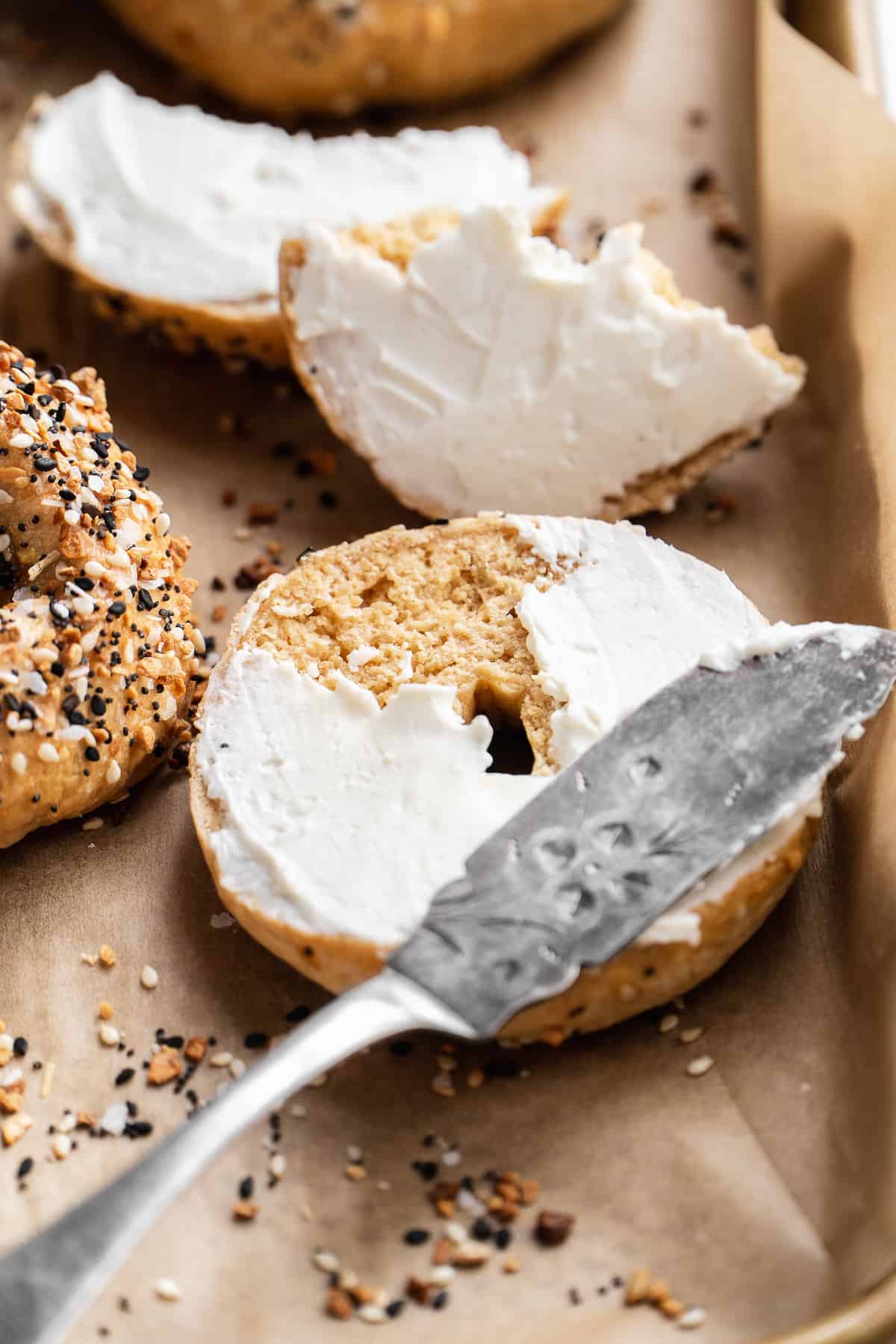 Gluten free everything bagel on a baking sheet with a knife spreading cream cheese on top.