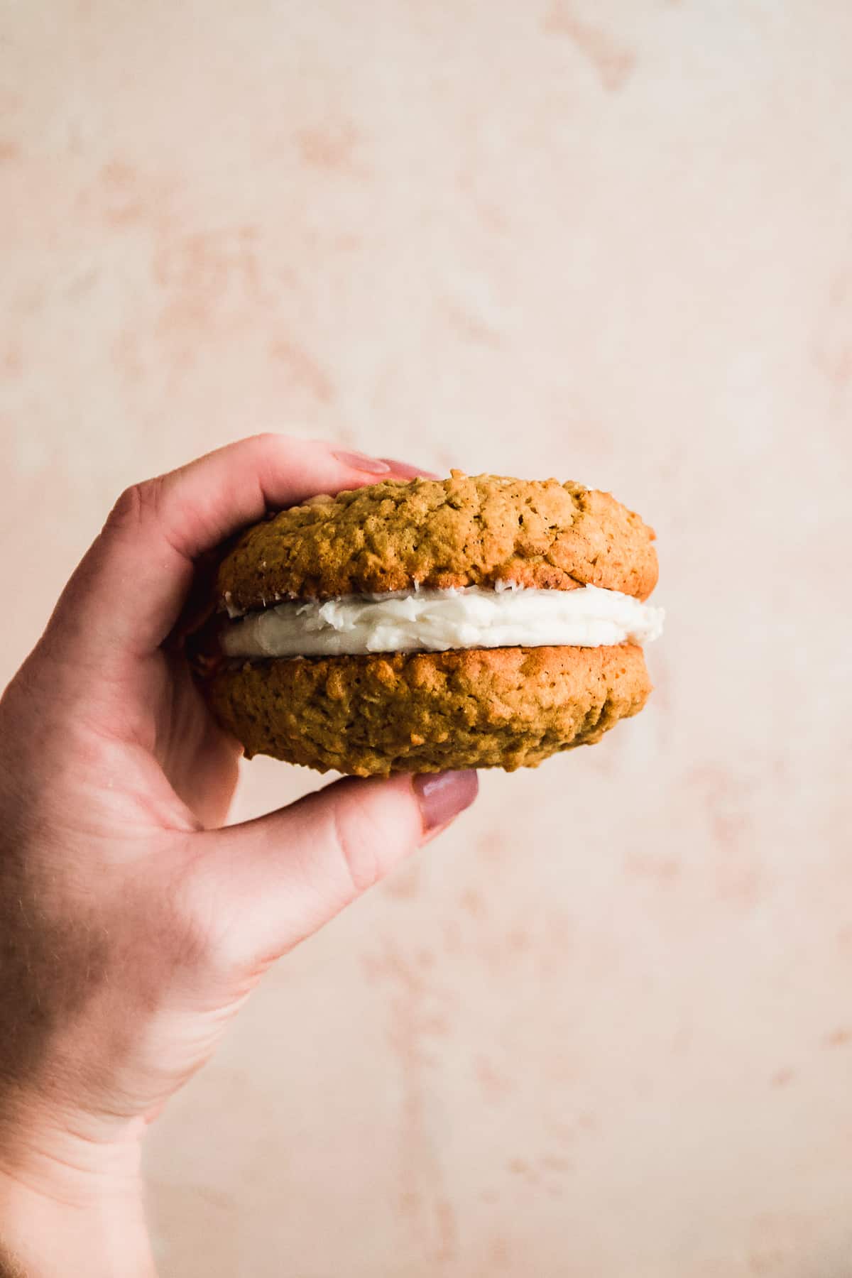 Hand holding up a gluten free oatmeal cream pie in front of a pink surface.