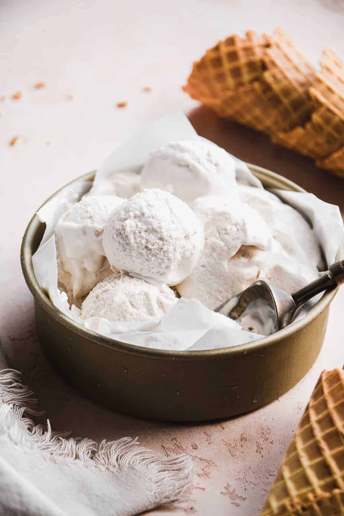 Scoops of vegan ice cream in a container with cones scattered on surface.