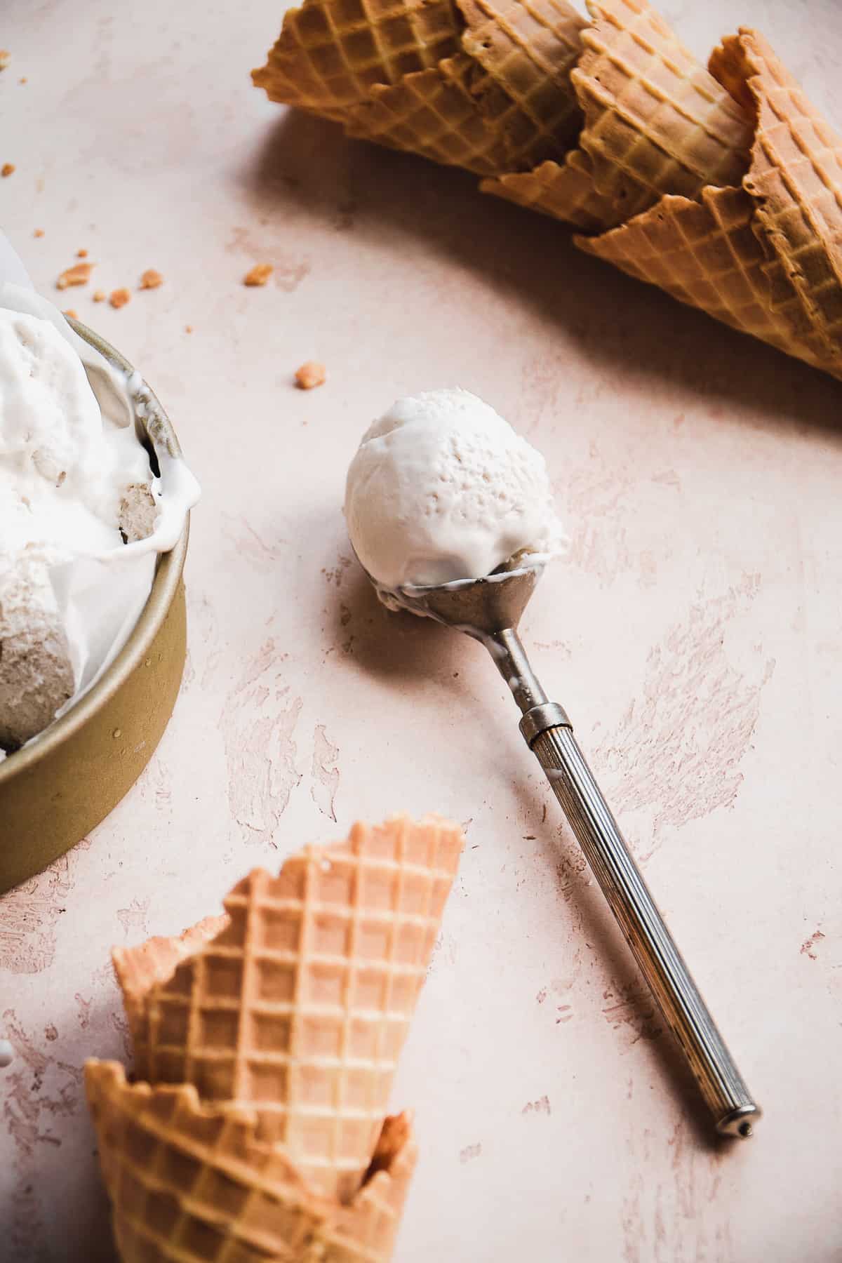 Ice cream scoop with dairy free vanilla ice cream and cones in the background.