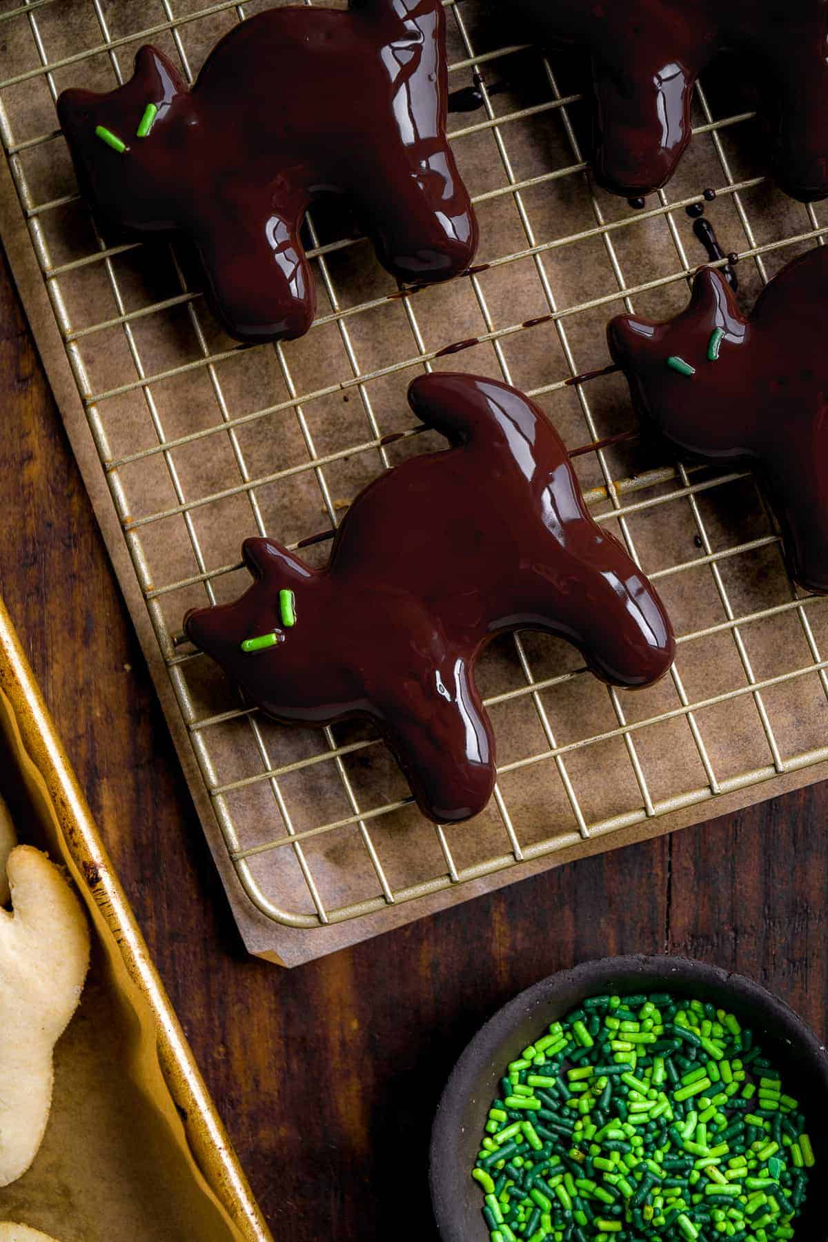 Cat cookie coated in melted chocolate with green sprinkles for eyes on a wire rack.