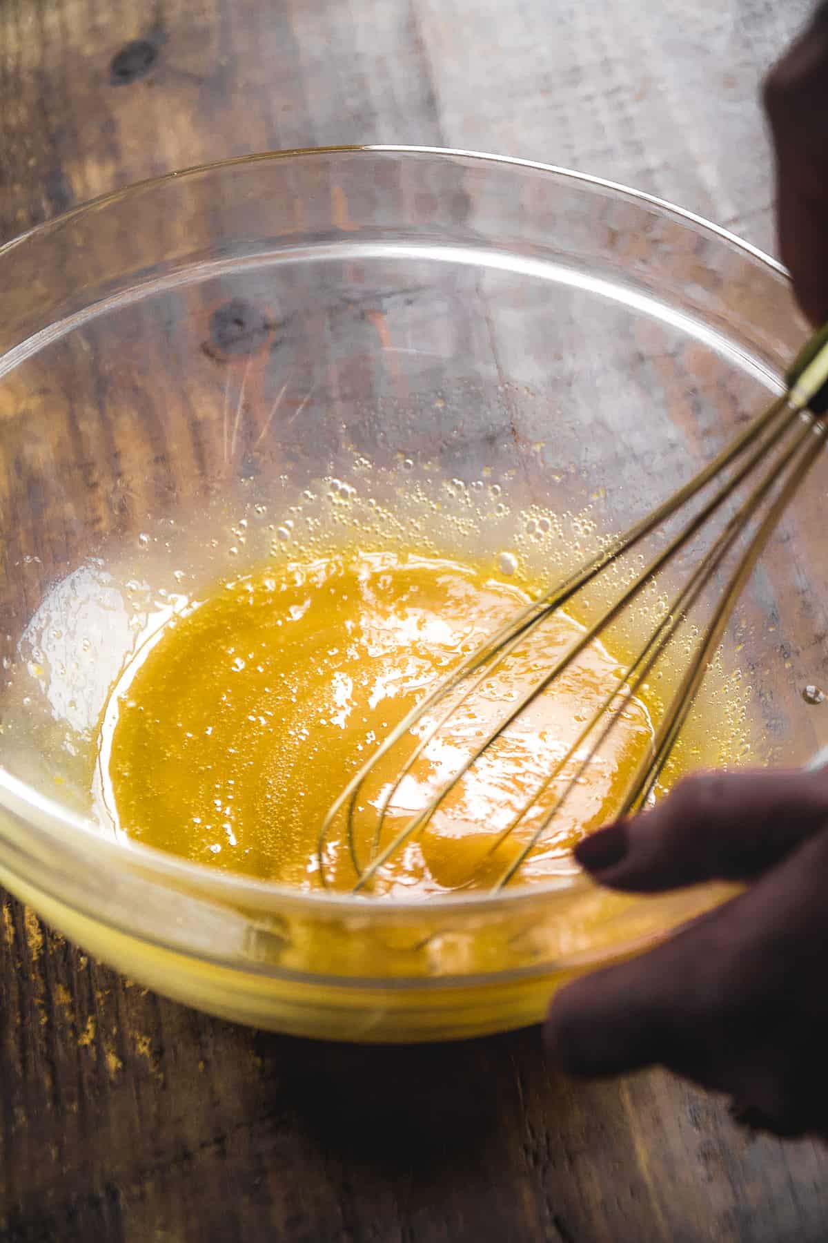 Person whisking wet cookie ingredients in a glass bowl on a wooden surface.