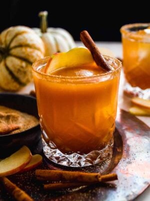 Pumpkin bourbon cocktail in short glass with cinnamon stick inside and pumpkins in background.