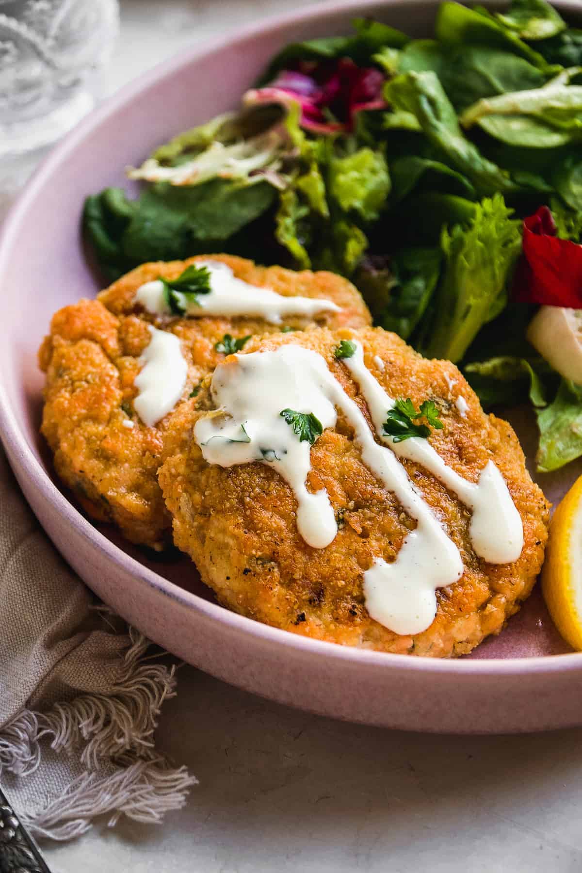 Two salmon cakes on a pink plate with sauce drizzled on top and a salad on the plate.