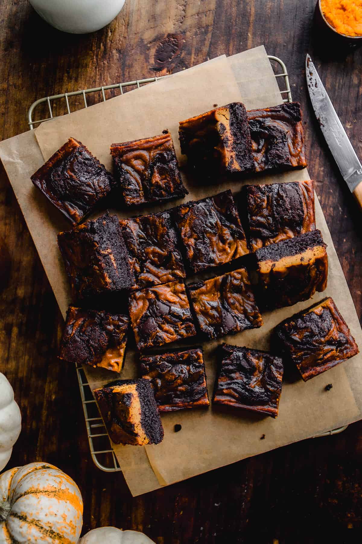 Brownies with pumpkin swirls scattered on brown parchment paper on wooden surface.