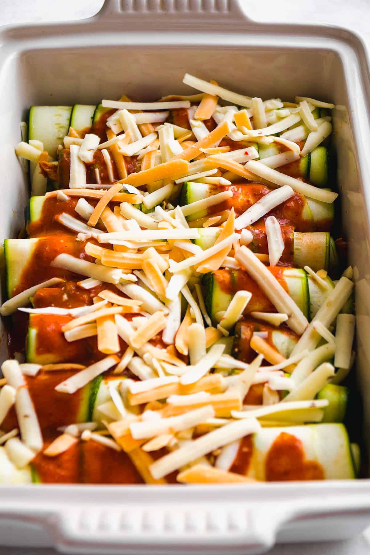 Baking dish with zucchini chicken roll ups topped with enchilada sauce and cheese about to be baked.