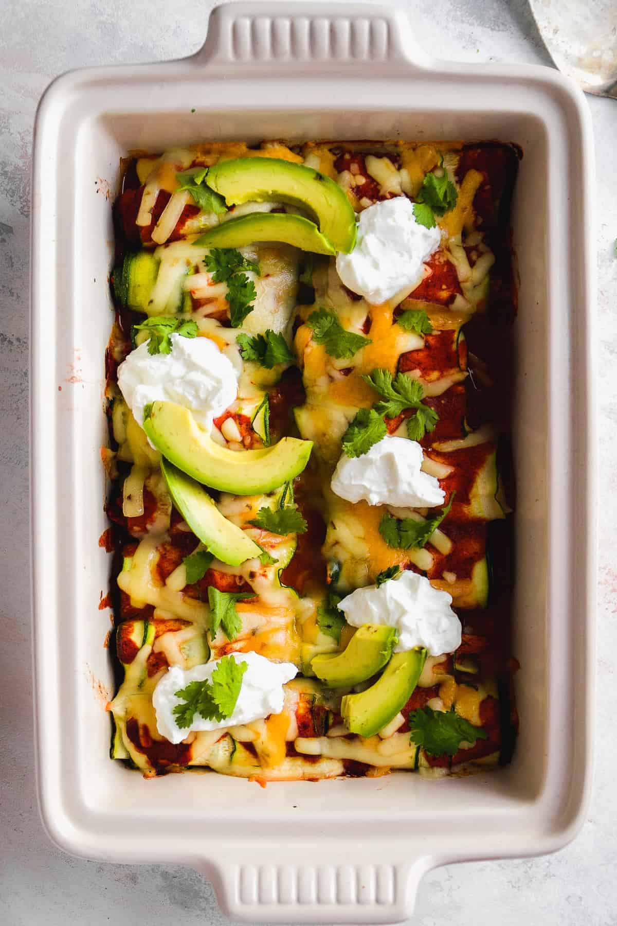 Overhead view of zucchini enchiladas baked in a white dish with avocados and sour cream on top.