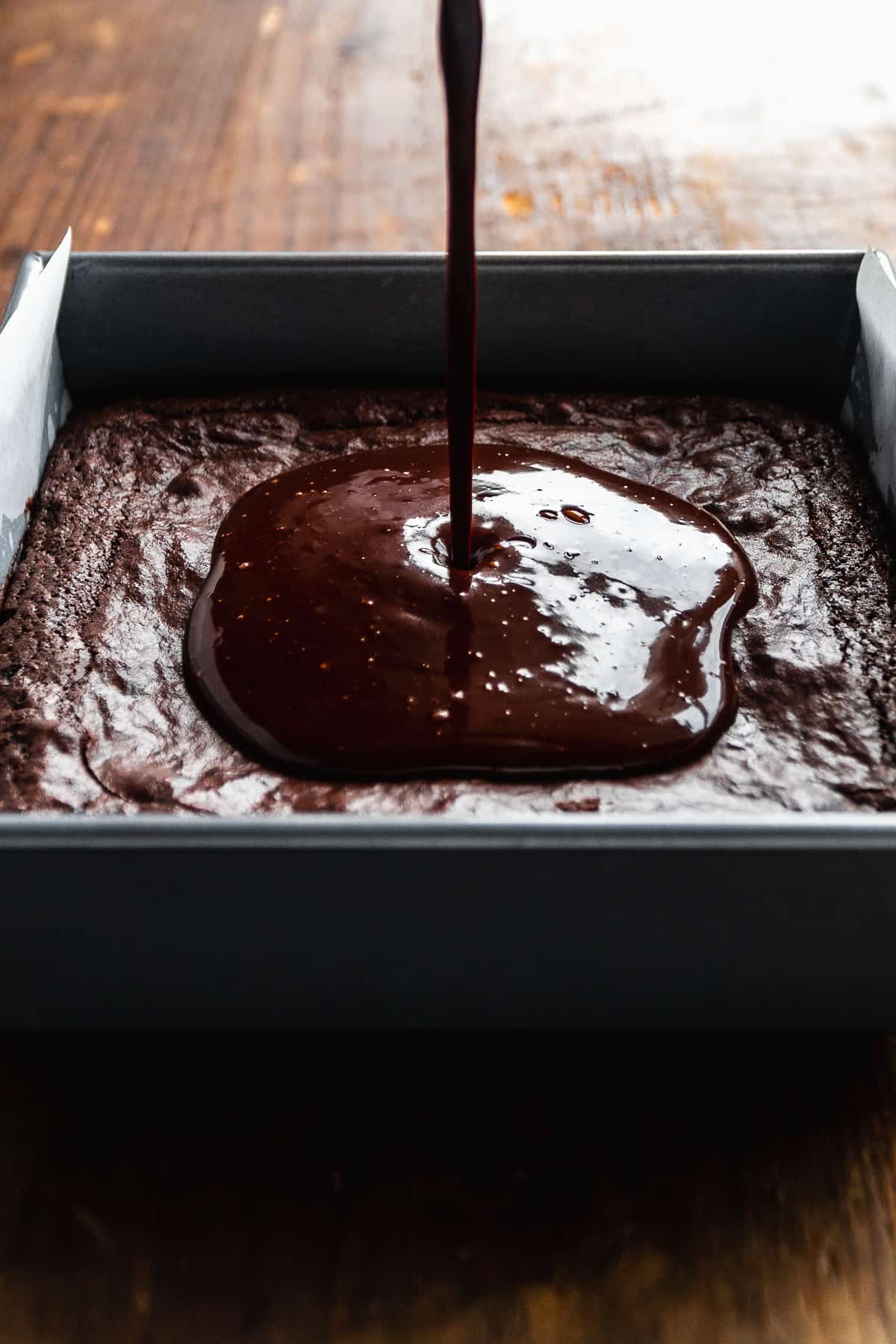 Chocolate being drizzled onto a pan of brownies on wooden surface.