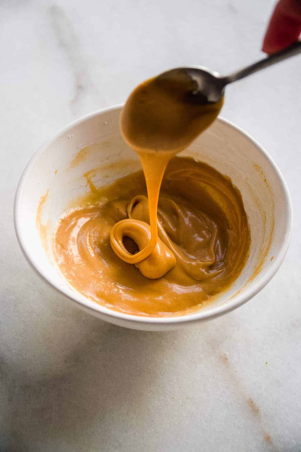 Spoon drizzling almond butter icing into a bowl on marble surface.