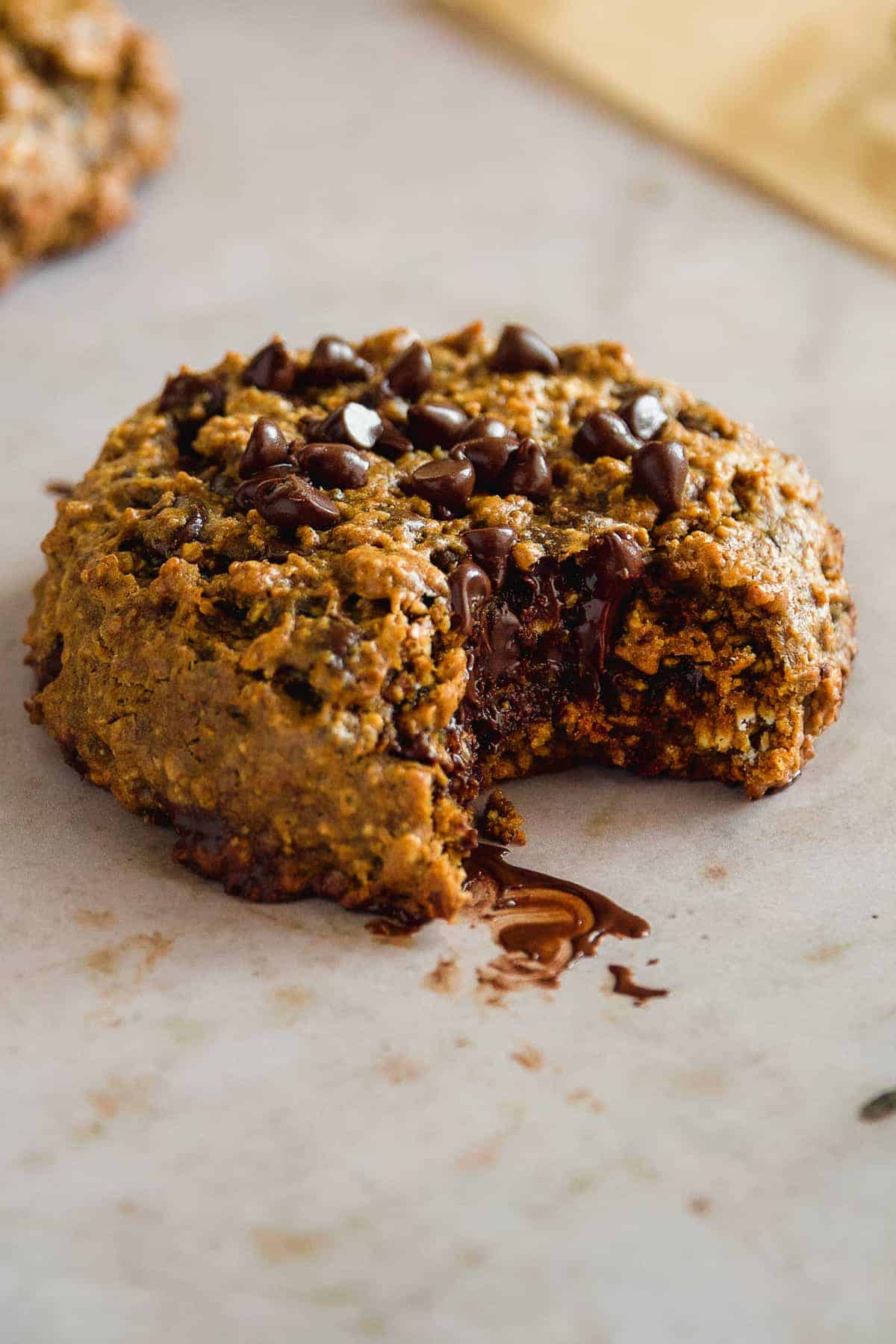 A pumpkin gluten free cookie with chocolate chips on top and a bite taken out of it on a tan surface.