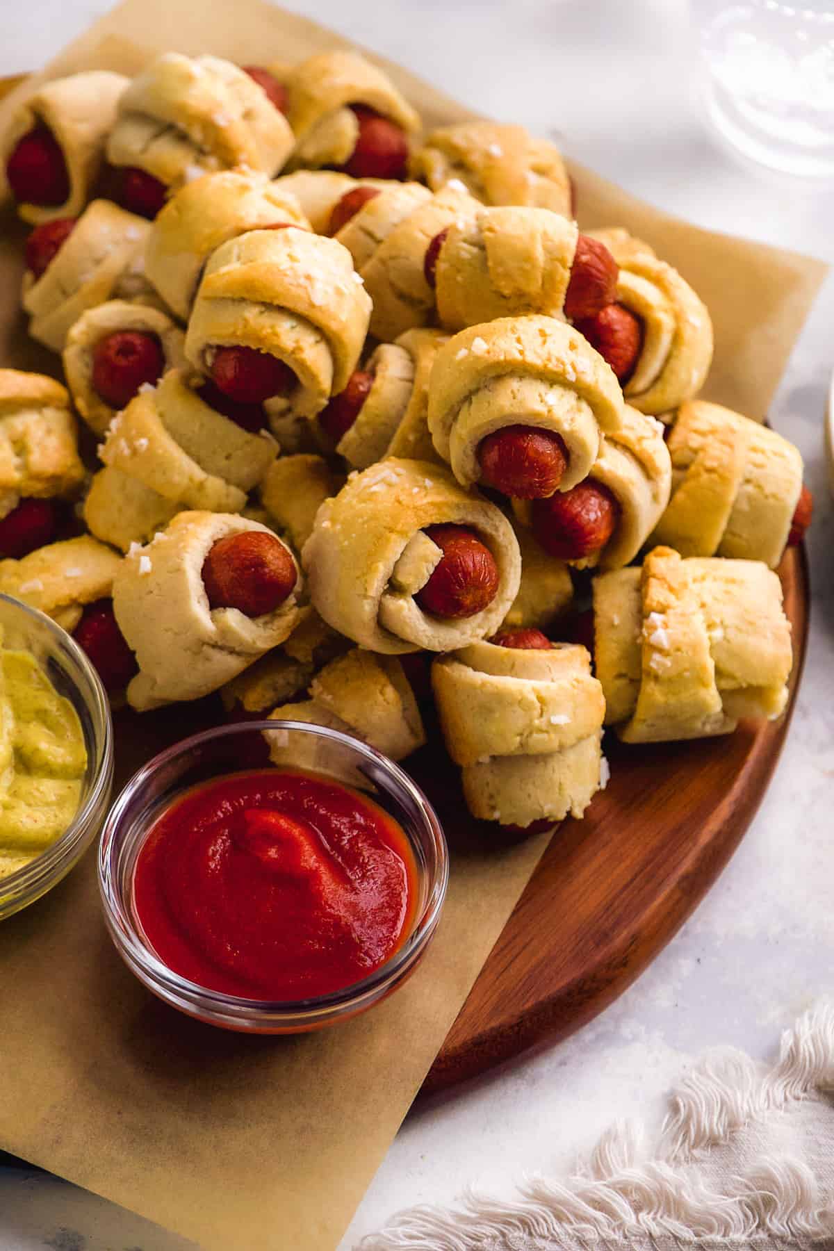 Gluten free pigs in a blanket piled up on a wooden platter with ketchup.