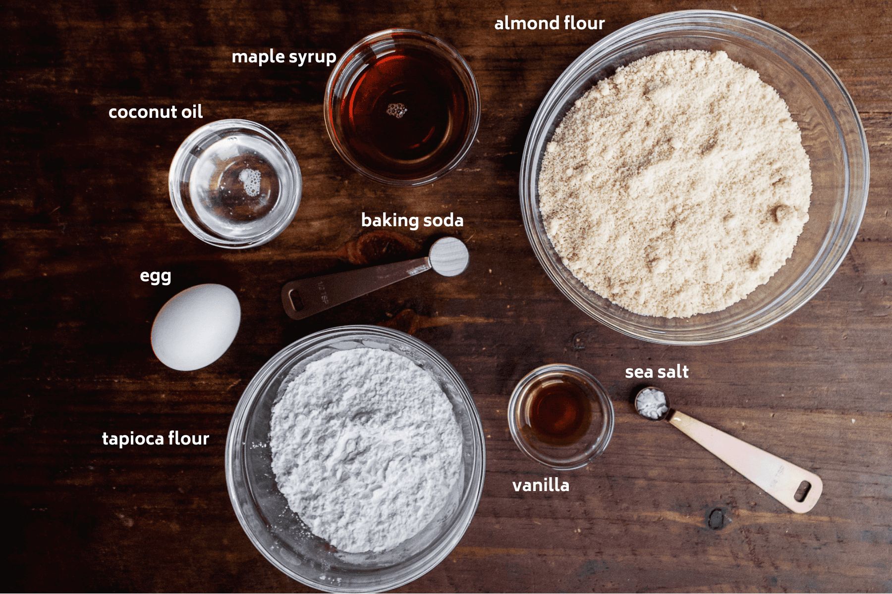 Top-down view of labeled baking ingredients for slice and bake cookies on a wooden surface: almond flour, tapioca flour, egg, coconut oil, maple syrup, vanilla, baking soda, and sea salt in bowls and measuring spoons.