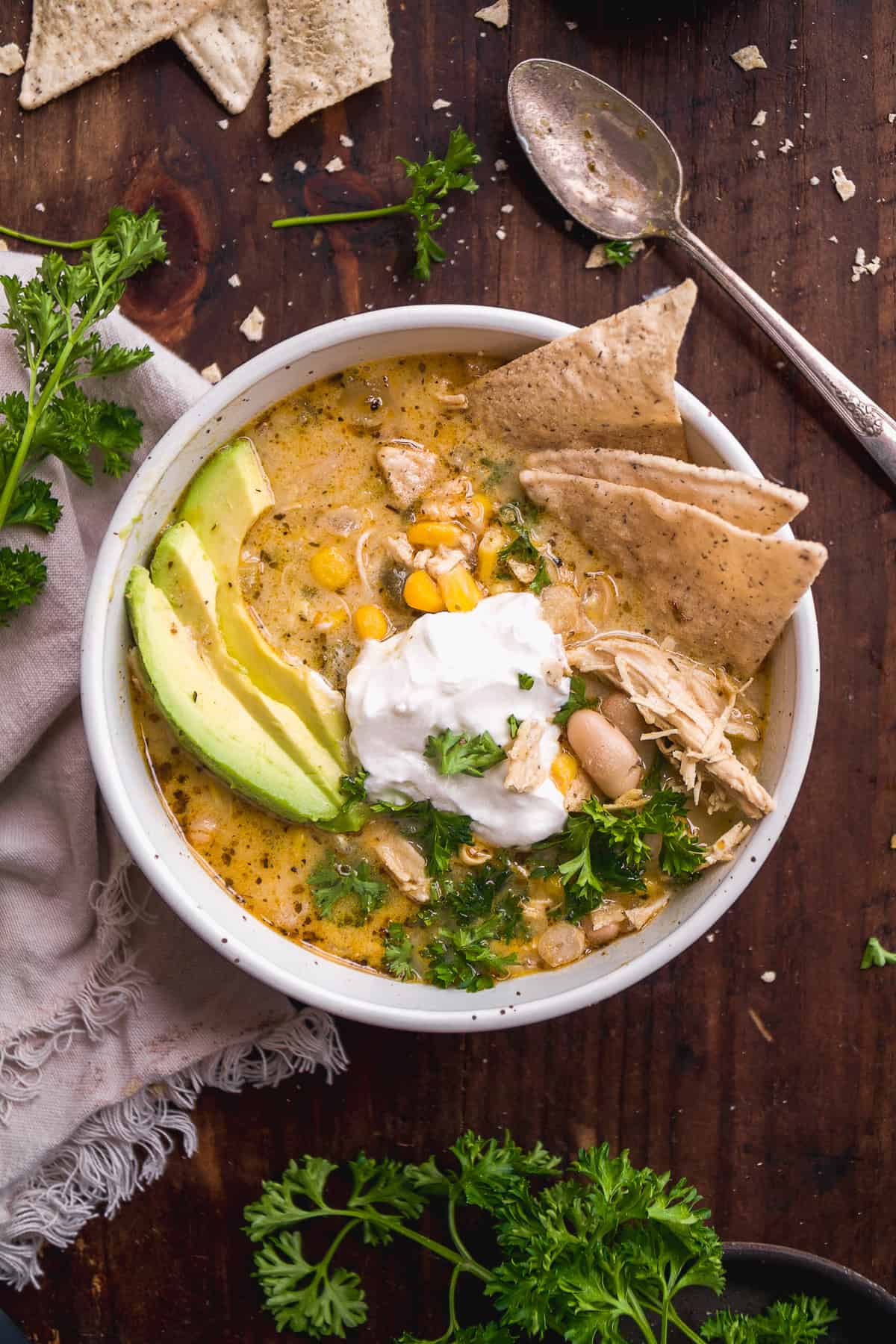 Overhead view of a bowl of chicken chili with chips and avocados inside on a wooden backdrop.