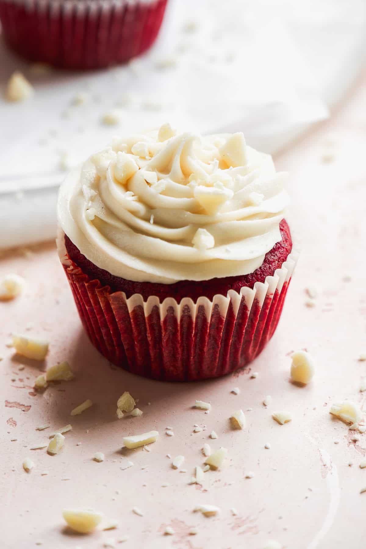 One gluten free red velvet cupcake with cream cheese frosting on top on a pink surface.