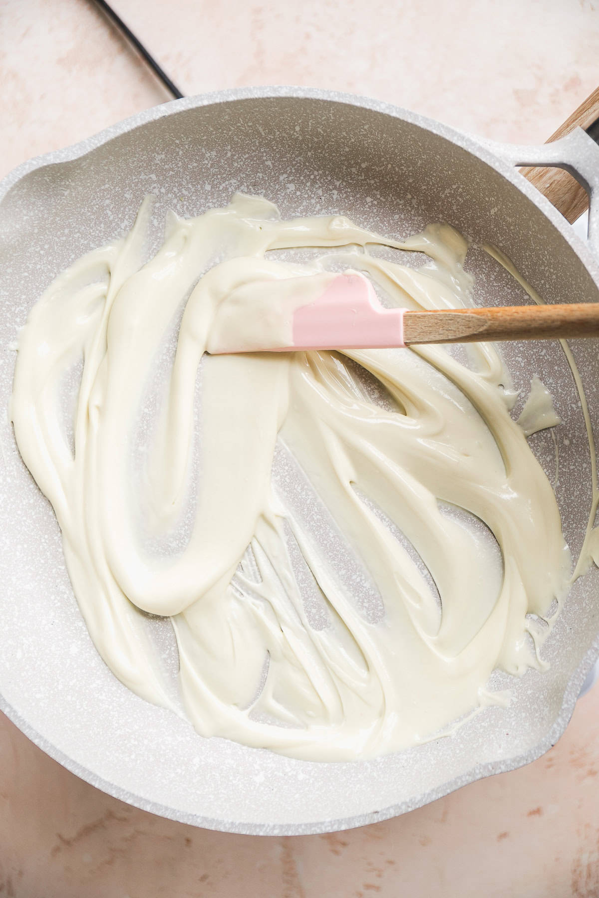 Melted white chocolate in a gray skillet with a pink spatula.