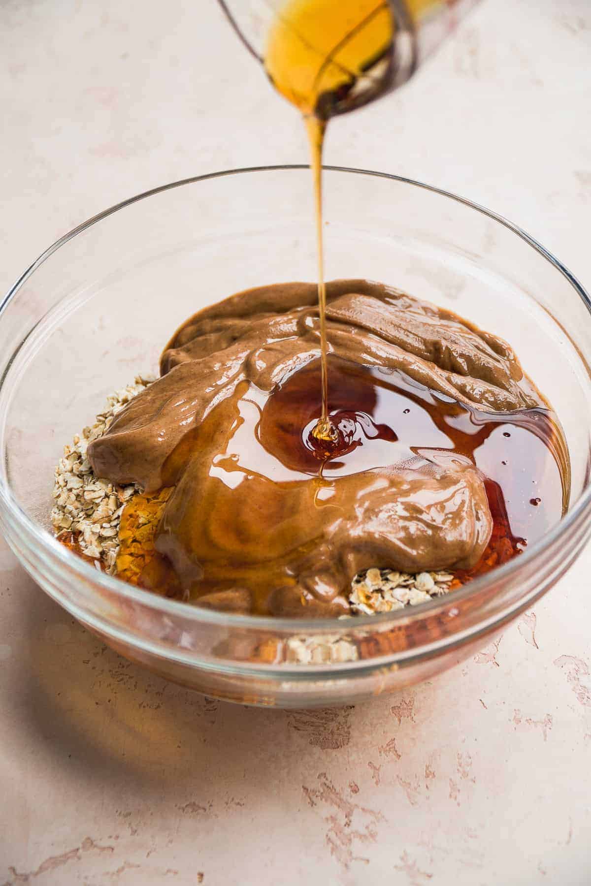 Honey being poured into a large glass mixing bowl with peanut butter and oats.