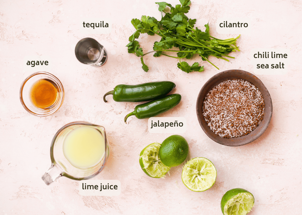 Ingredients for a jalapeño margarita are arranged on a light surface: cilantro, two jalapeños, chili lime sea salt in a bowl, squeezed limes, lime juice, agave, and a jigger of tequila, each labeled with text.