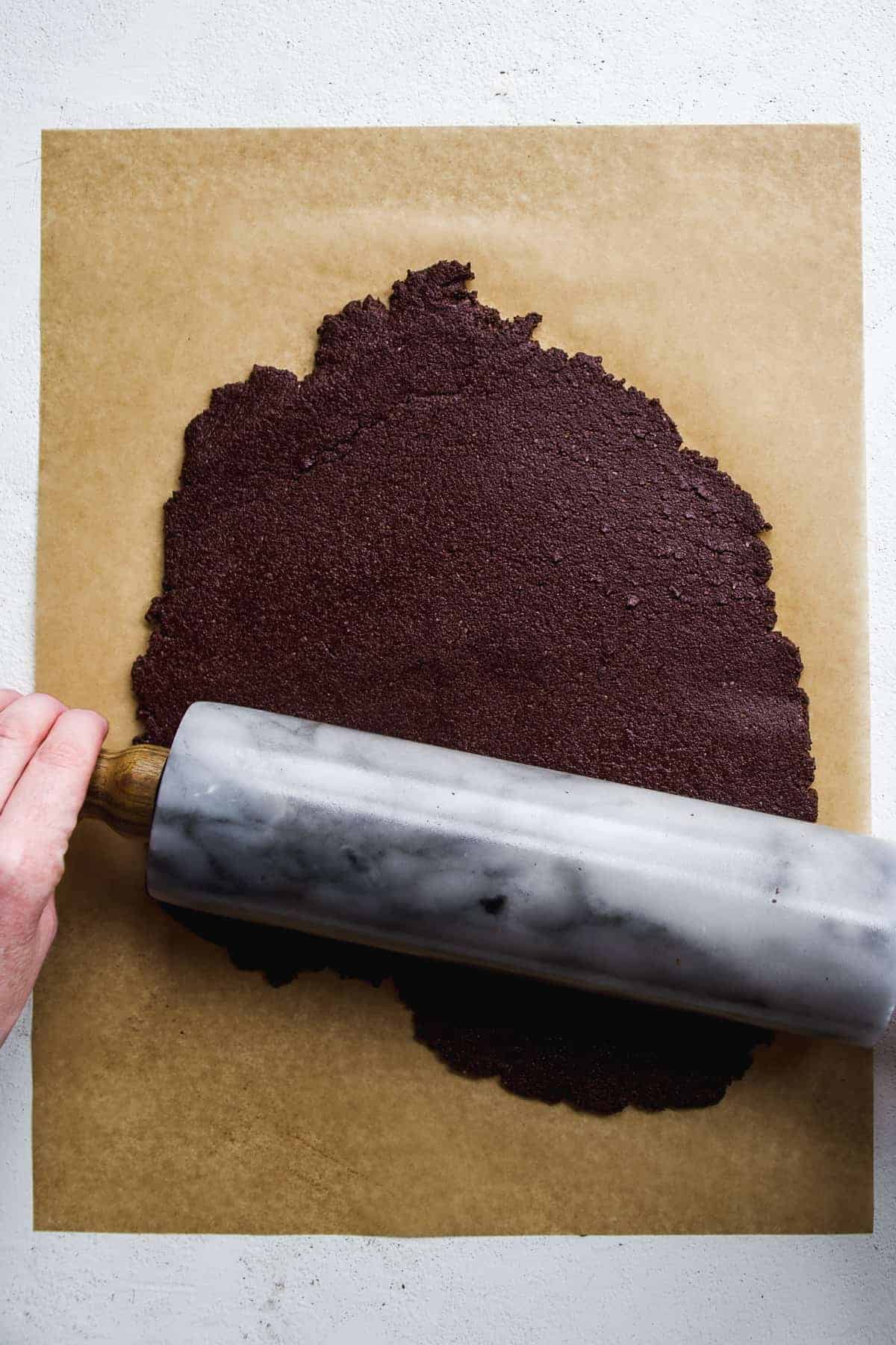 Person rolling out chocolate cookie dough on a piece of parchment paper.