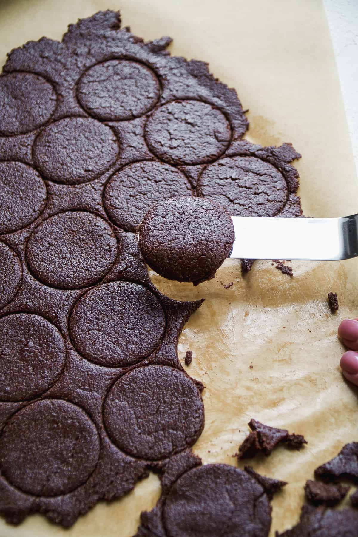 Circles being cut out of chocolate cookie dough and a metal spatula lifting one up.