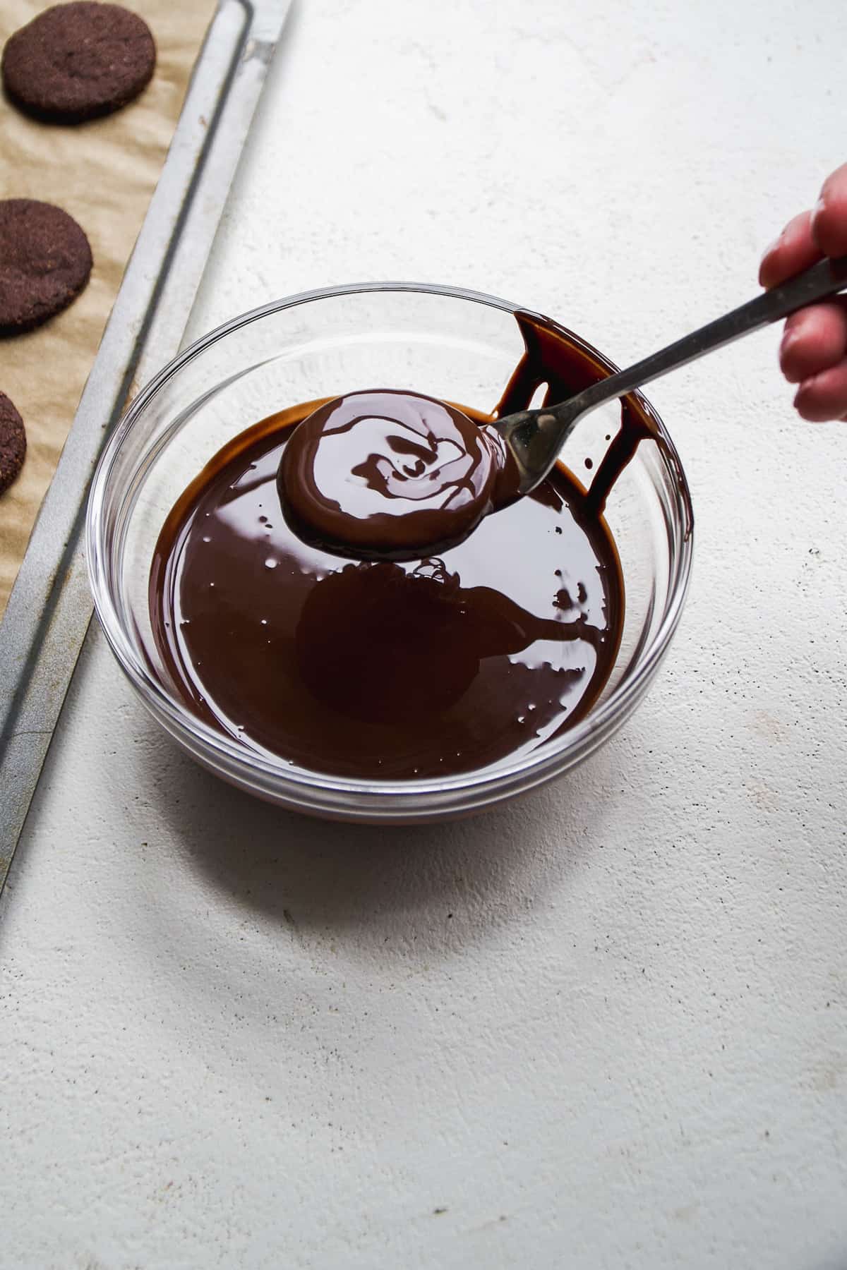 Thin chocolate cookies being dunked in a bowl of melted chocolate with a fork.