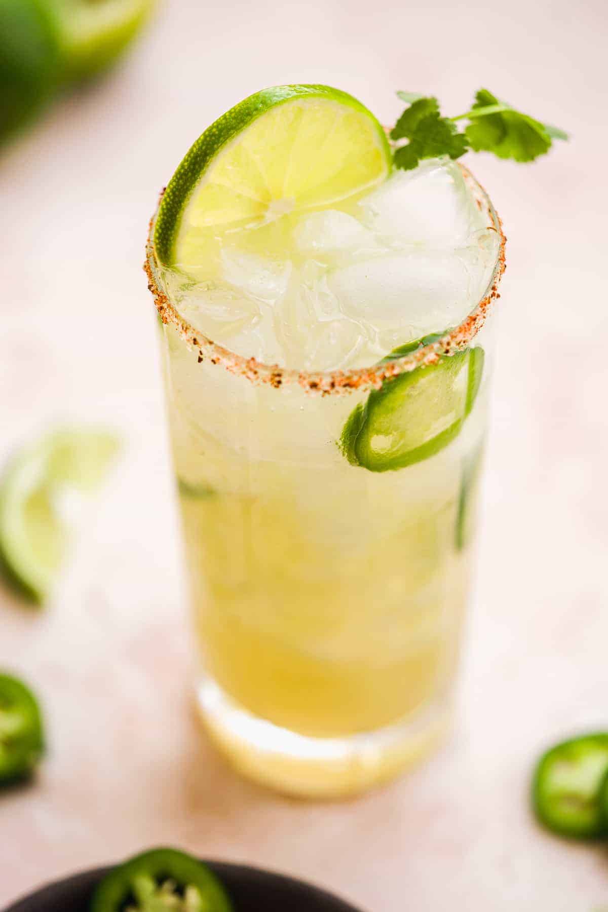 Tall glass of margarita with lime slice on top and a jalapeño and orange salted rim.