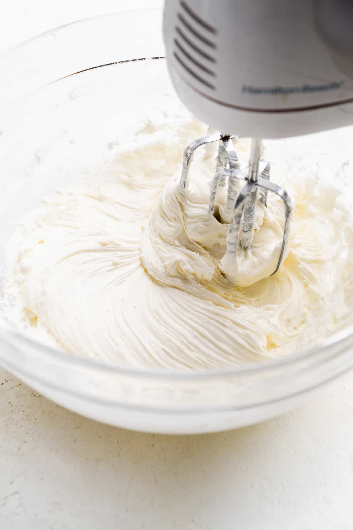 Cream cheese icing being mixed in a clear bowl with a hand mixer.