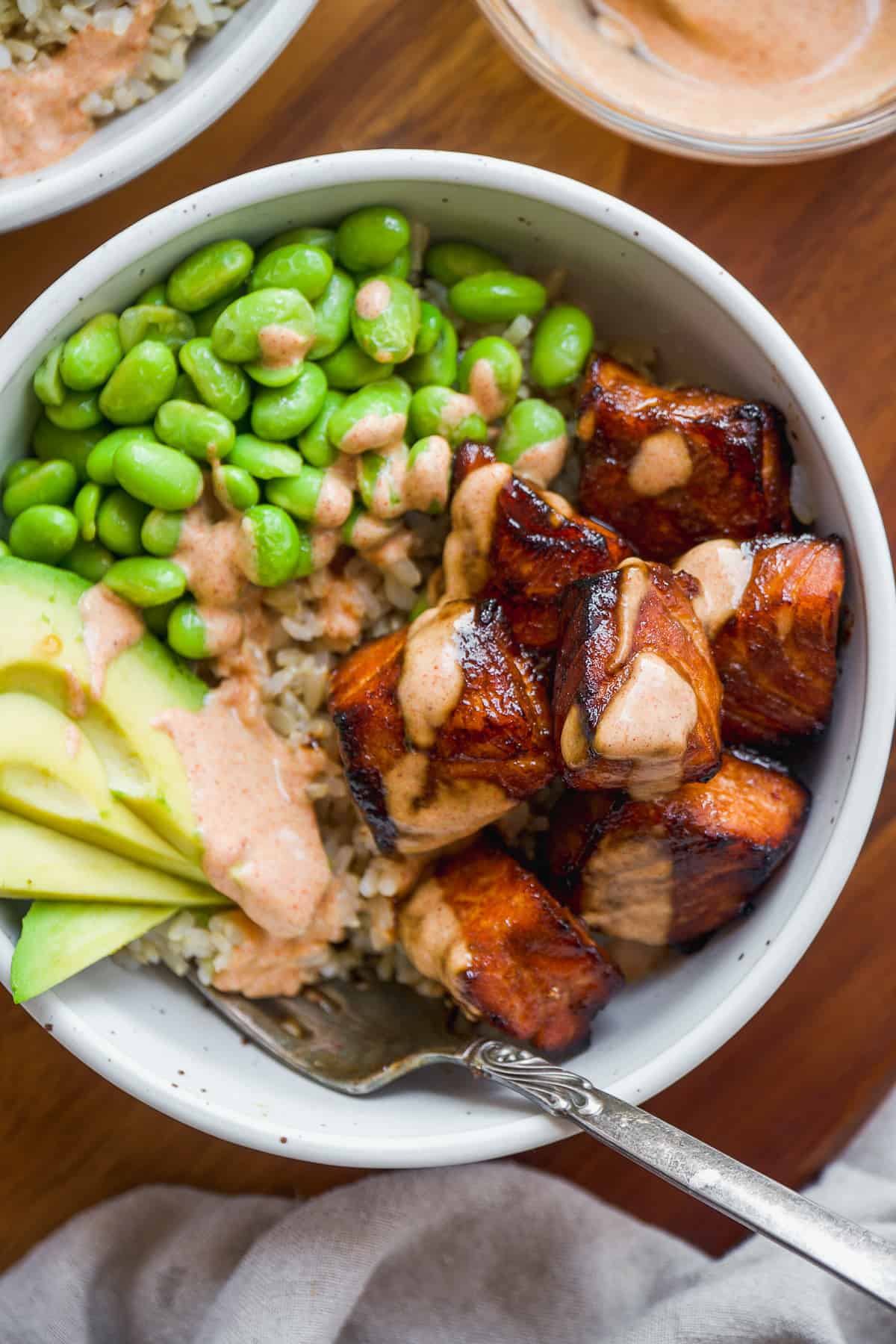 Teriyaki salmon bites in a bowl with brown rice, edamame, and avocado with orange sauce on top.