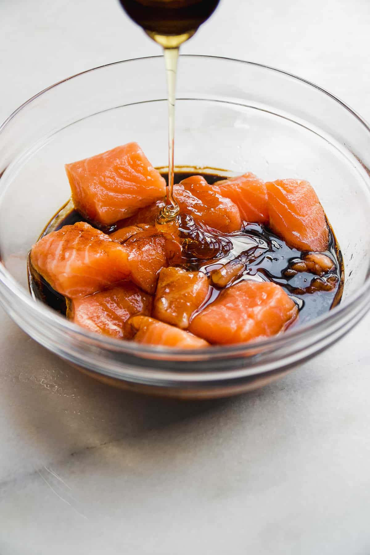 Honey being poured into a bowl with salmon cubes and teriyaki sauce.