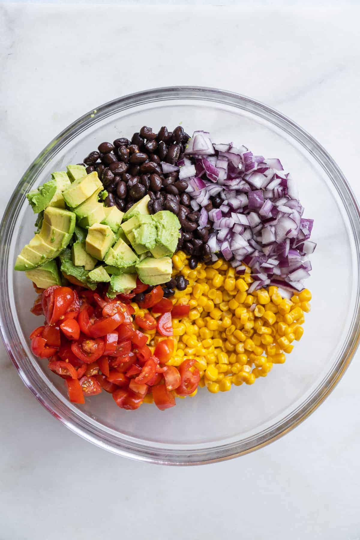 Clear bowl on a marble surface with corn salad ingredients about to be mixed in.