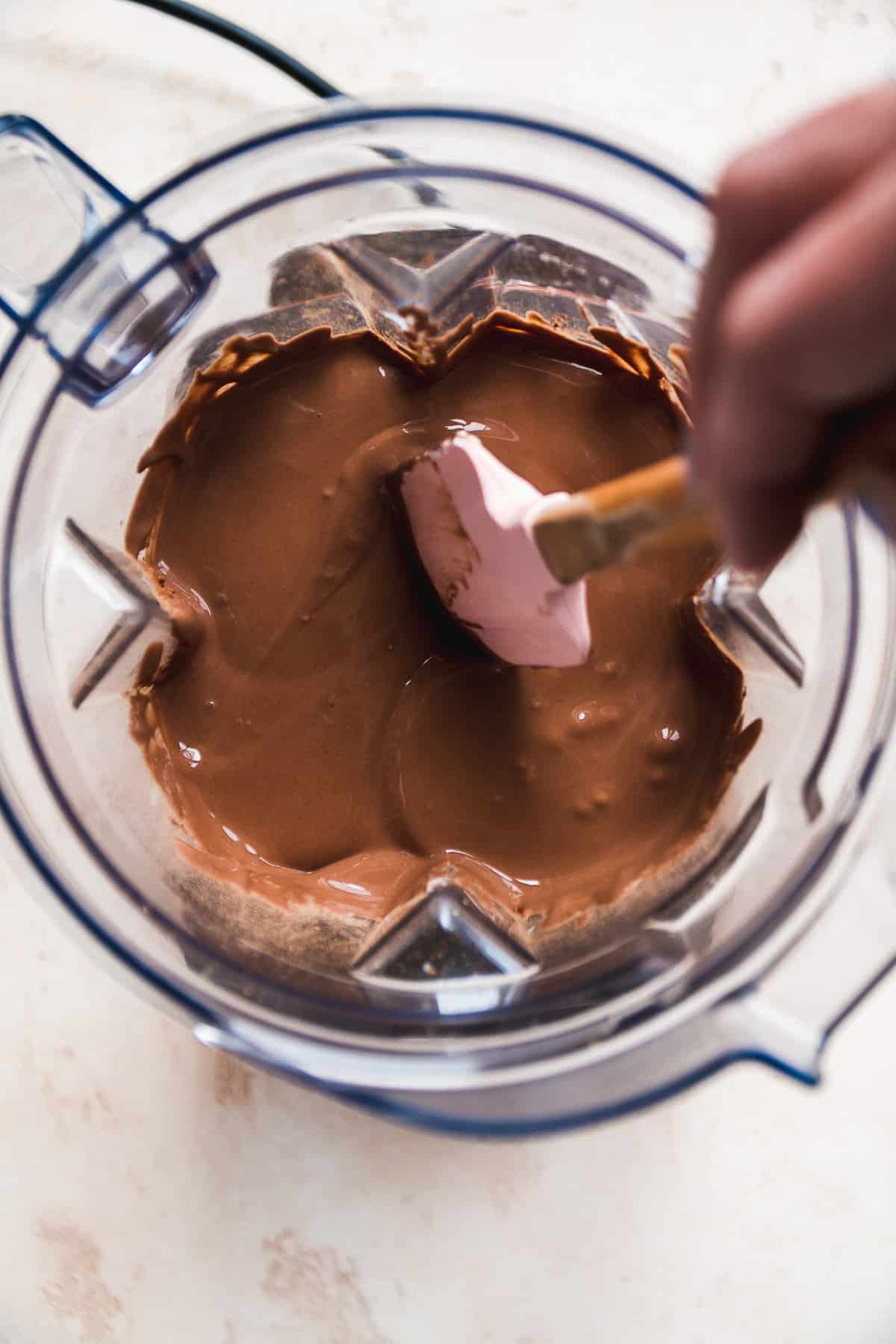 Person stirring chocolate greek yogurt in a blender with a spatula.