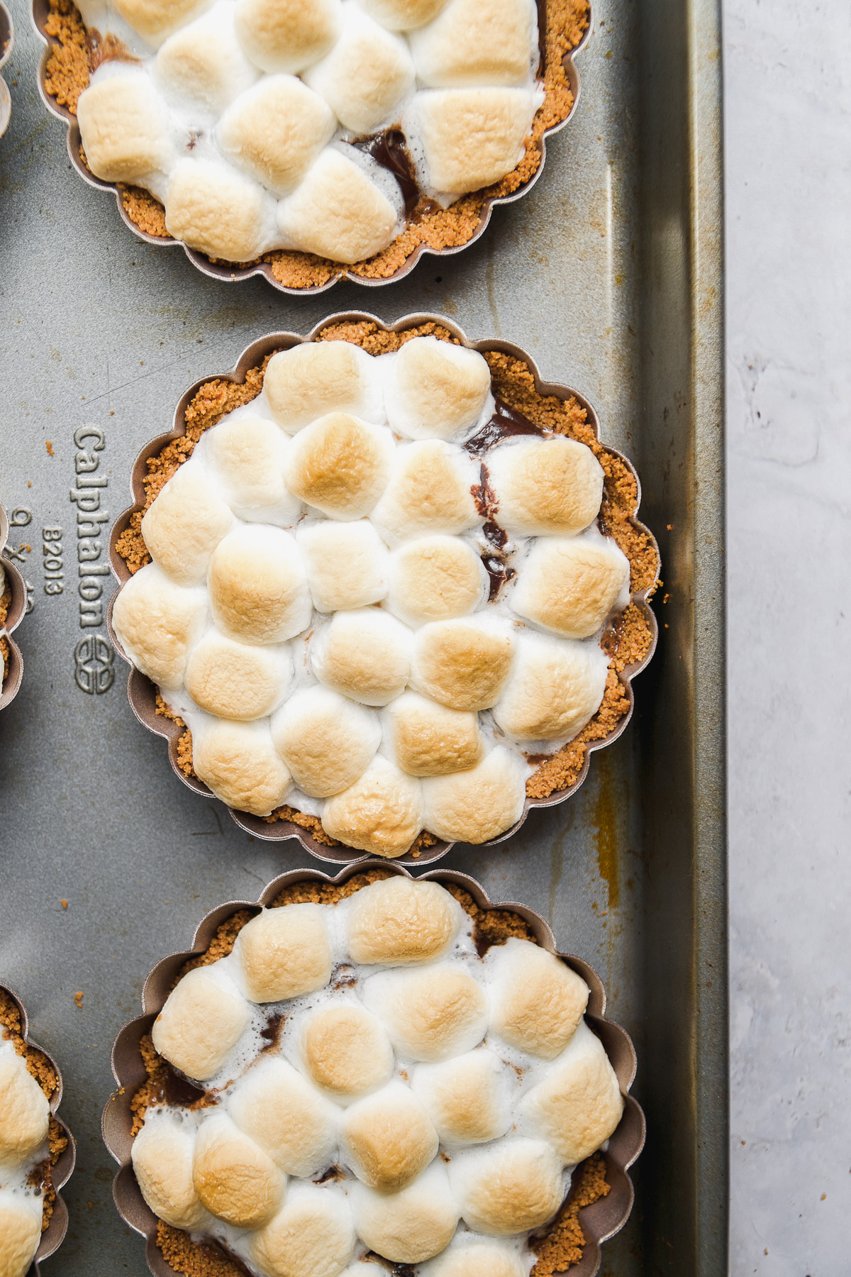 Mini s'mores pies on a baking pan with toasted marshmallows on top.