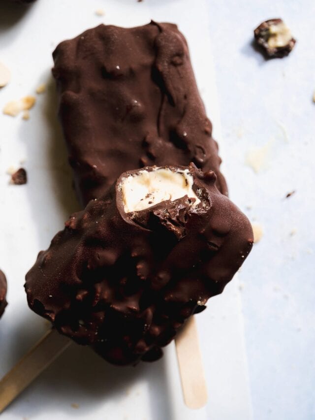 Two chocolate-coated ice cream bars on wooden sticks, one with a bite taken out, revealing creamy vanilla ice cream inside. Chocolate crumbs are scattered on the white surface around them.