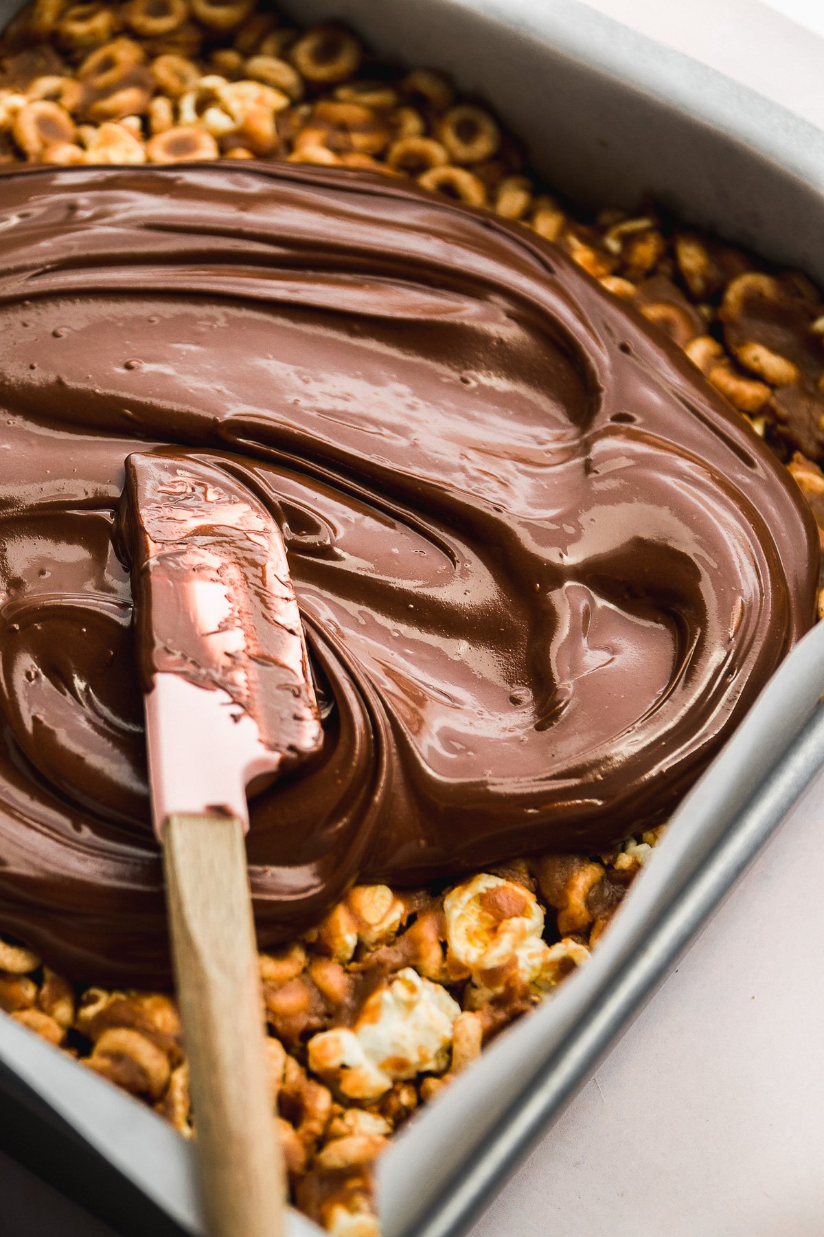 Rubber spatula spreading melted chocolate over peanut butter cheerio bars.