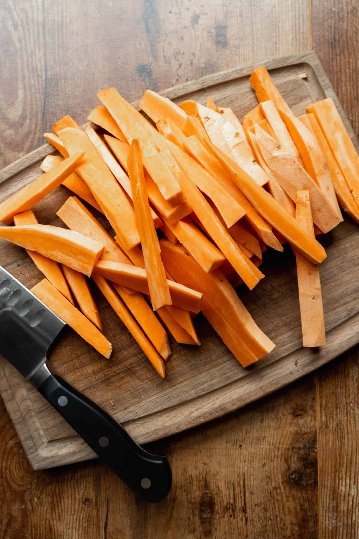 Sweet potatoes cut into fries on a wooden cutting board with a knife.