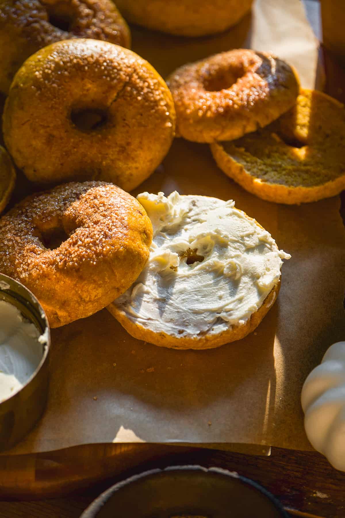 Pumpkin bagels scattered on parchment paper with cream cheese spread on one.