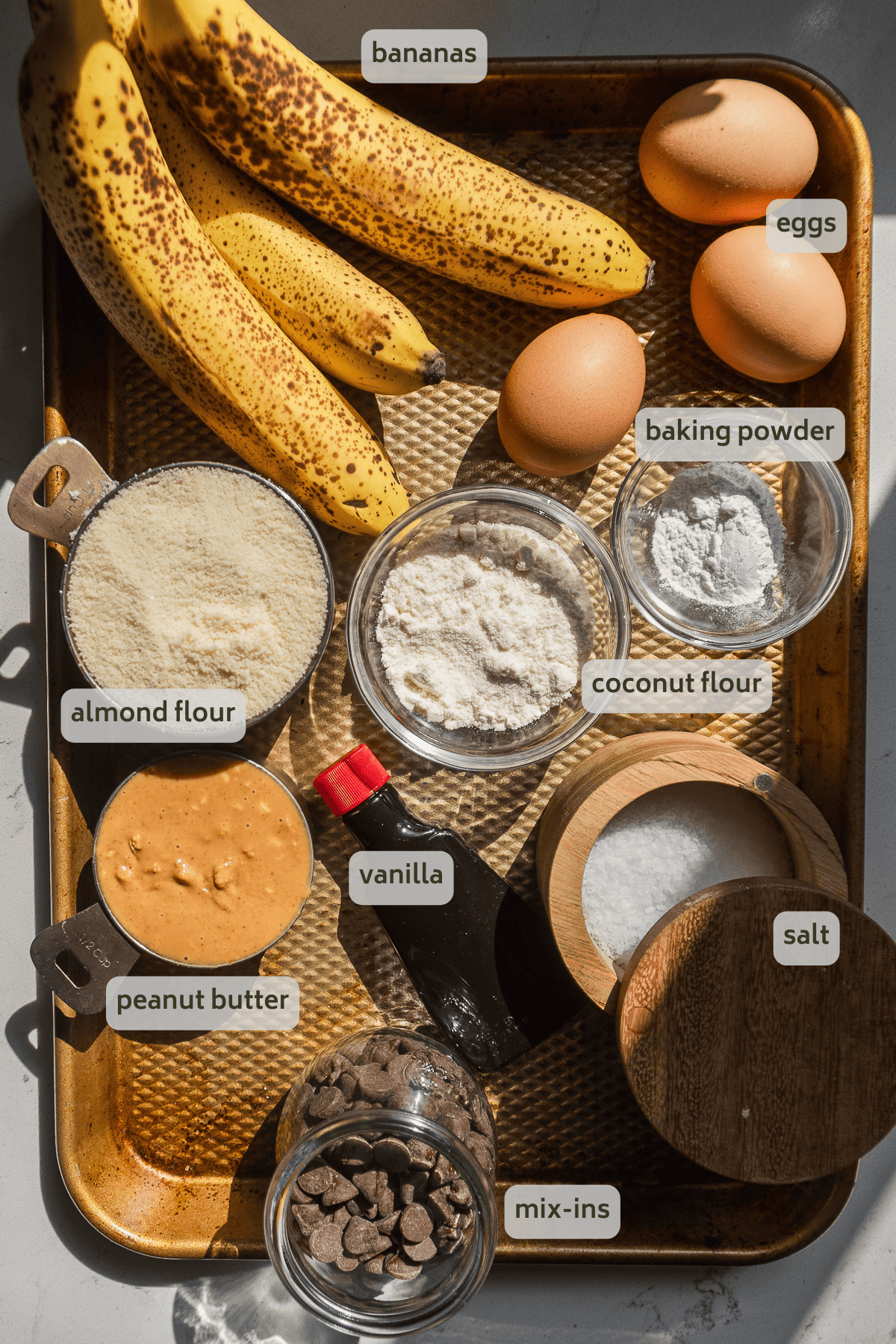 Banana bread ingredients with labels on a gold baking sheet.