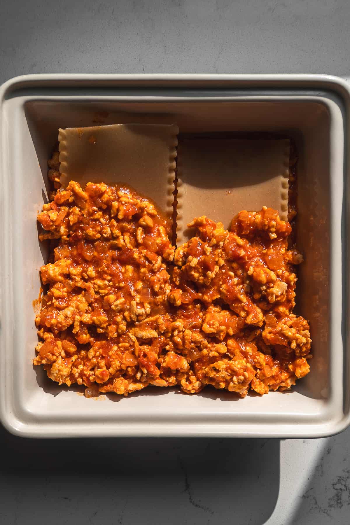 Lasagna being assembled in a square baking dish.