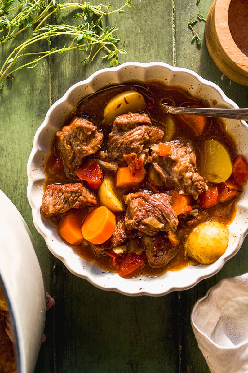 Beef stew with baby potatoes and carrots in a scalloped bowl on a green surface.